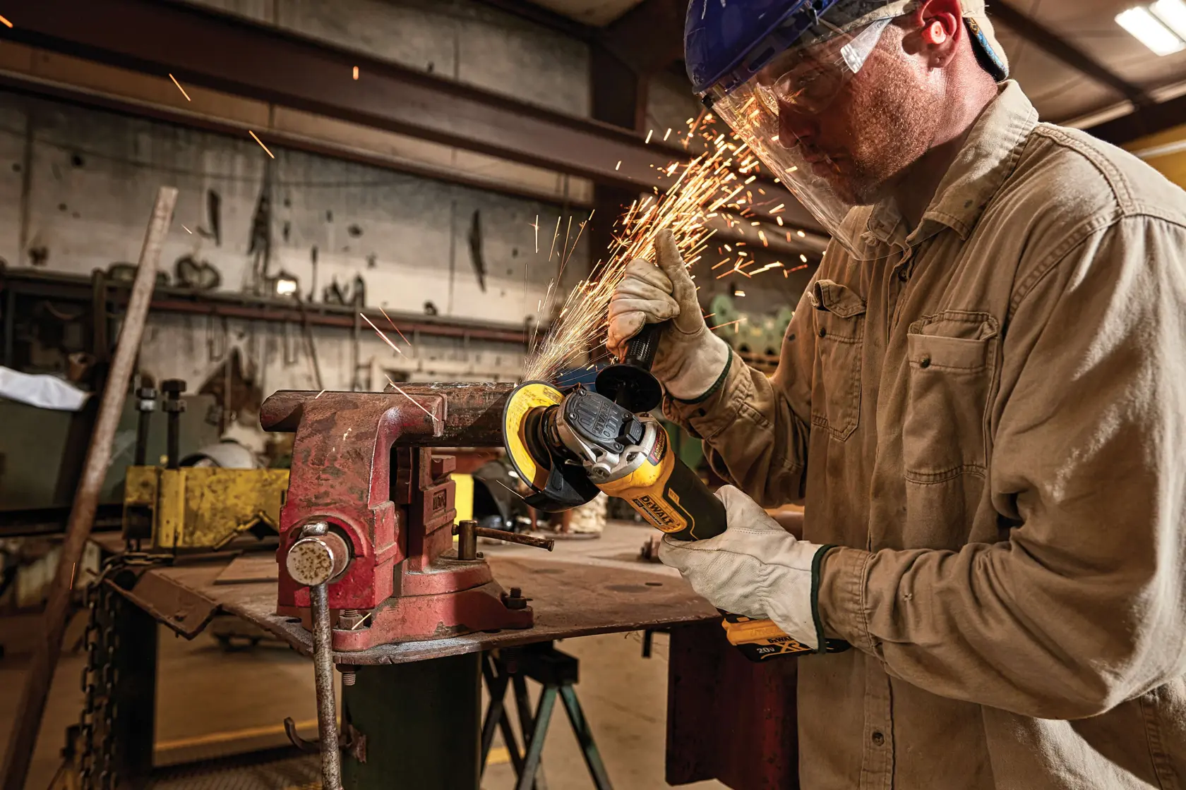 A worker uses a DEWALT cordless angle grinder to cut metal, producing sparks, in an industrial workshop setting. The worker is wearing gloves and safety gear.