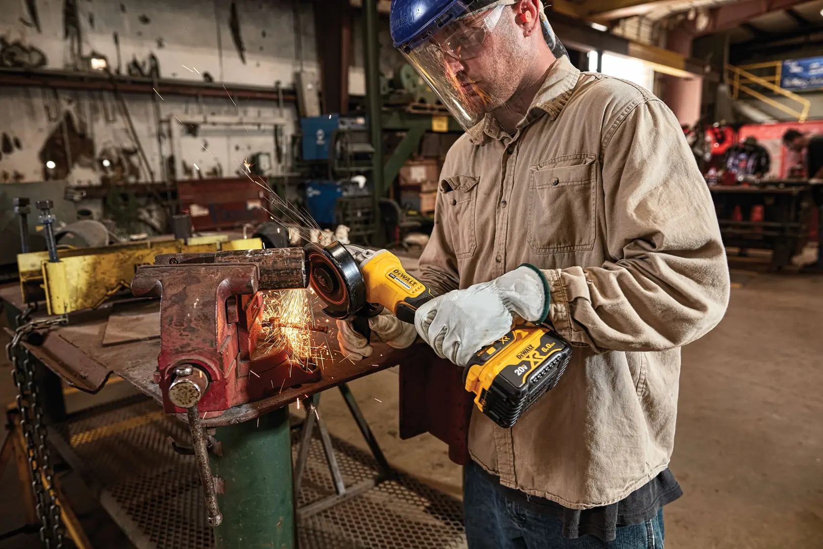 A person wearing protective gloves and a jacket is using a DEWALT cordless angle grinder to work on a piece of metal clamped in a vice, creating sparks in an industrial workshop setting.