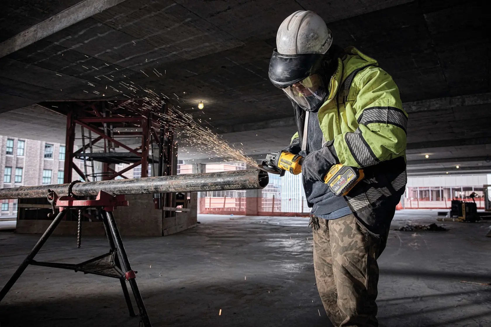Person wearing safety gear uses a DEWALT DCG414B cordless angle grinder to cut a metal pipe, producing sparks, inside a construction site.