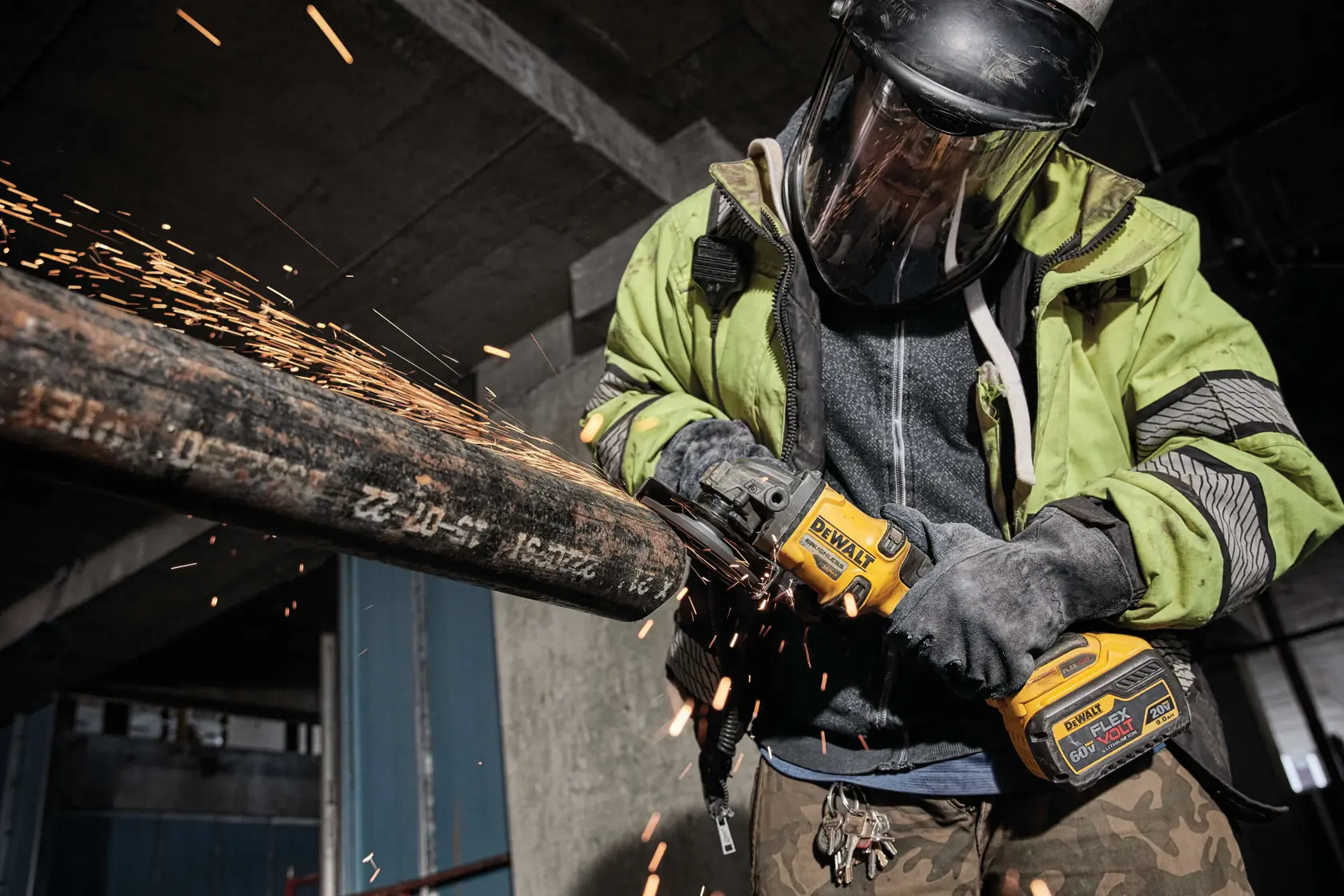 A worker wearing protective gear is using a DEWALT cordless angle grinder (model DCG414B) to cut a large metal pipe, producing sparks in an industrial setting.