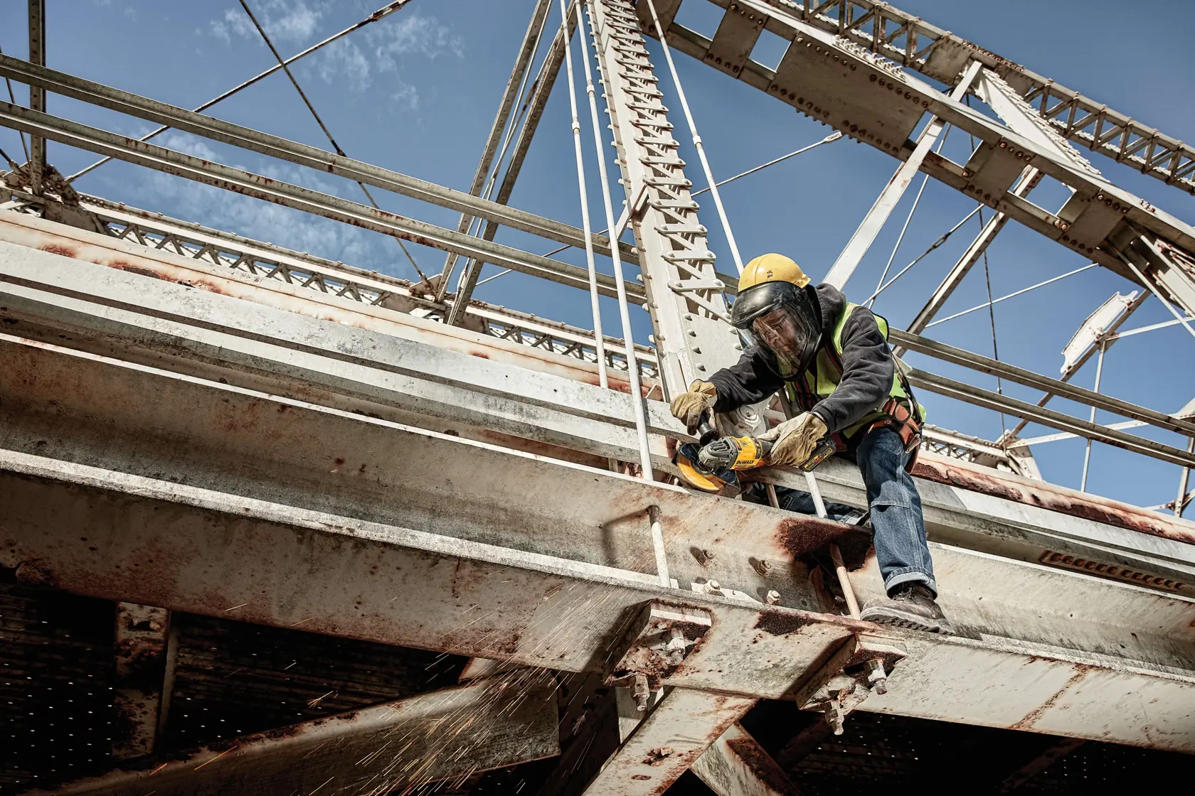 A construction worker wearing safety gear uses a DEWALT power tool to cut through a metal beam on a large steel bridge. Sparks are visible where the tool makes contact with the metal.