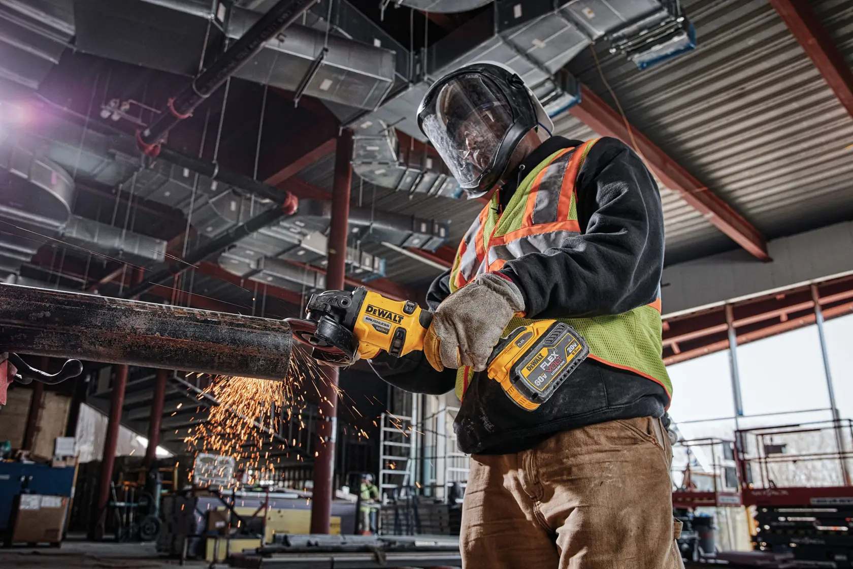 A worker wearing protective gear uses a DEWALT DCG414B cordless grinder to cut metal pipe, producing sparks, inside an industrial building under construction.