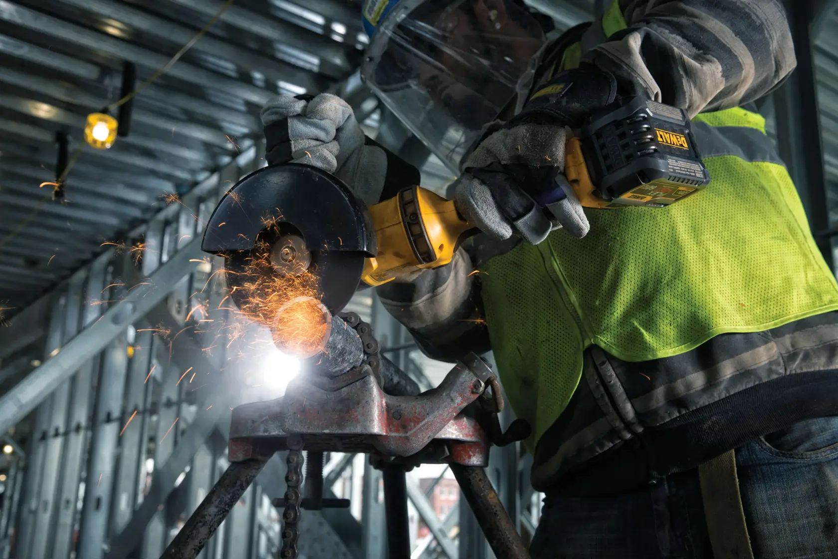 A close-up of a person wearing protective gloves and a safety vest using a DEWALT cordless angle grinder to cut metal, with sparks flying, inside an industrial construction site.