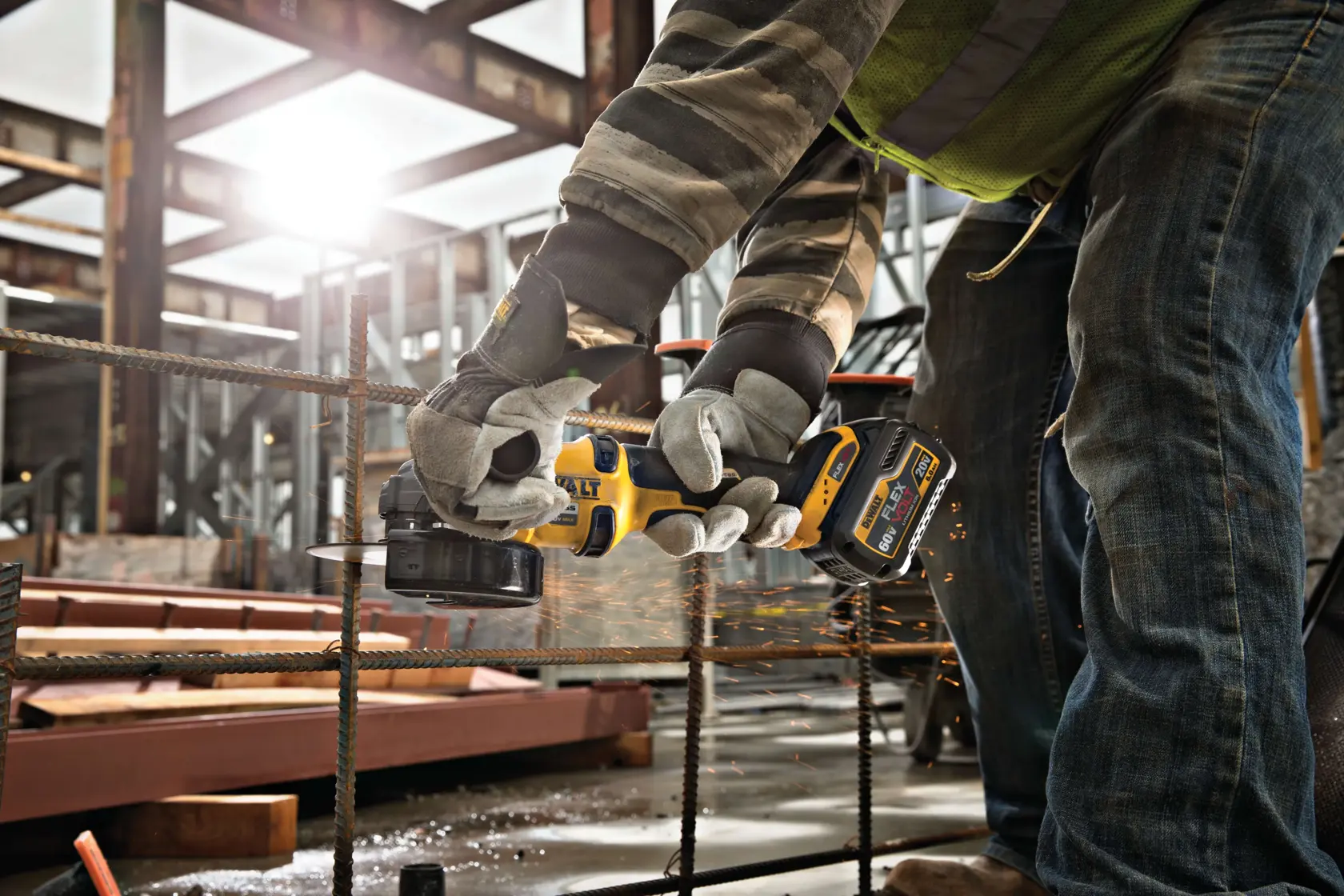 A person wearing gloves and safety gear uses a DEWALT cordless angle grinder to cut through metal rebar on a construction site, with sparks flying from the cutting process.