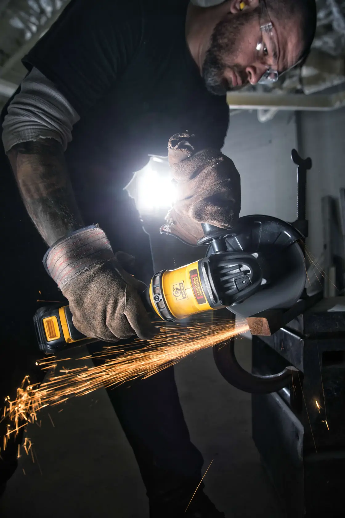 A person wearing protective gloves and dark clothing uses a DEWALT cordless angle grinder to cut metal, creating bright sparks. The face of the person is blurred for privacy. The workspace is industrial, with strong backlighting.