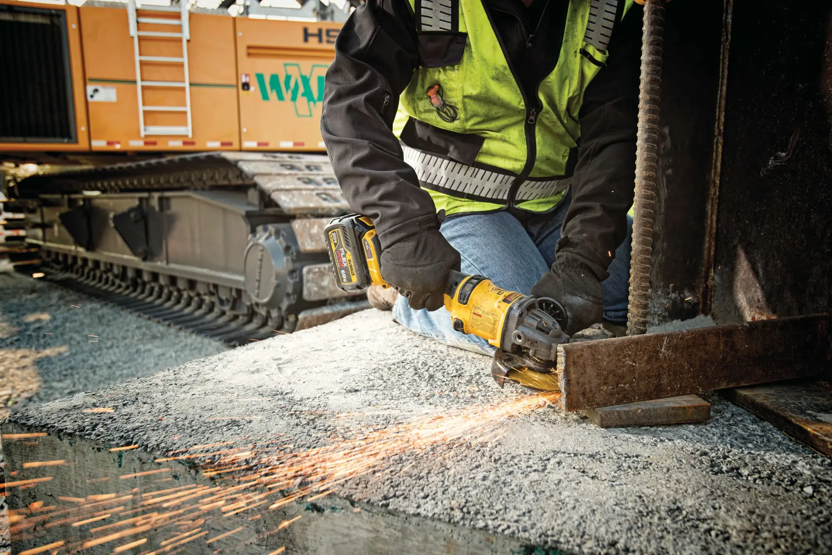 A person wearing a safety vest and gloves is using a DEWALT cordless angle grinder to cut through a piece of metal at a construction site. Sparks are flying from the metal, and there is construction machinery in the background.