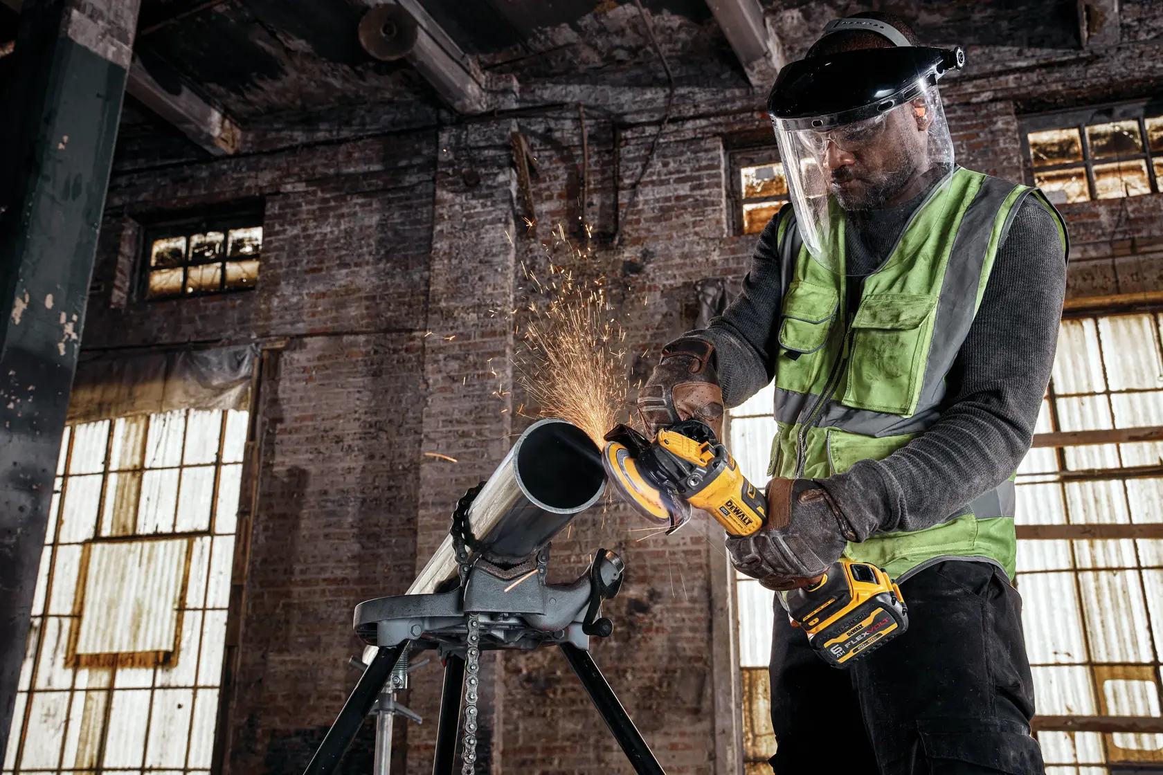 A person wearing a safety vest, gloves, and face shield is using a DEWALT cordless angle grinder on a metal pipe, with sparks flying, in an industrial workshop setting.