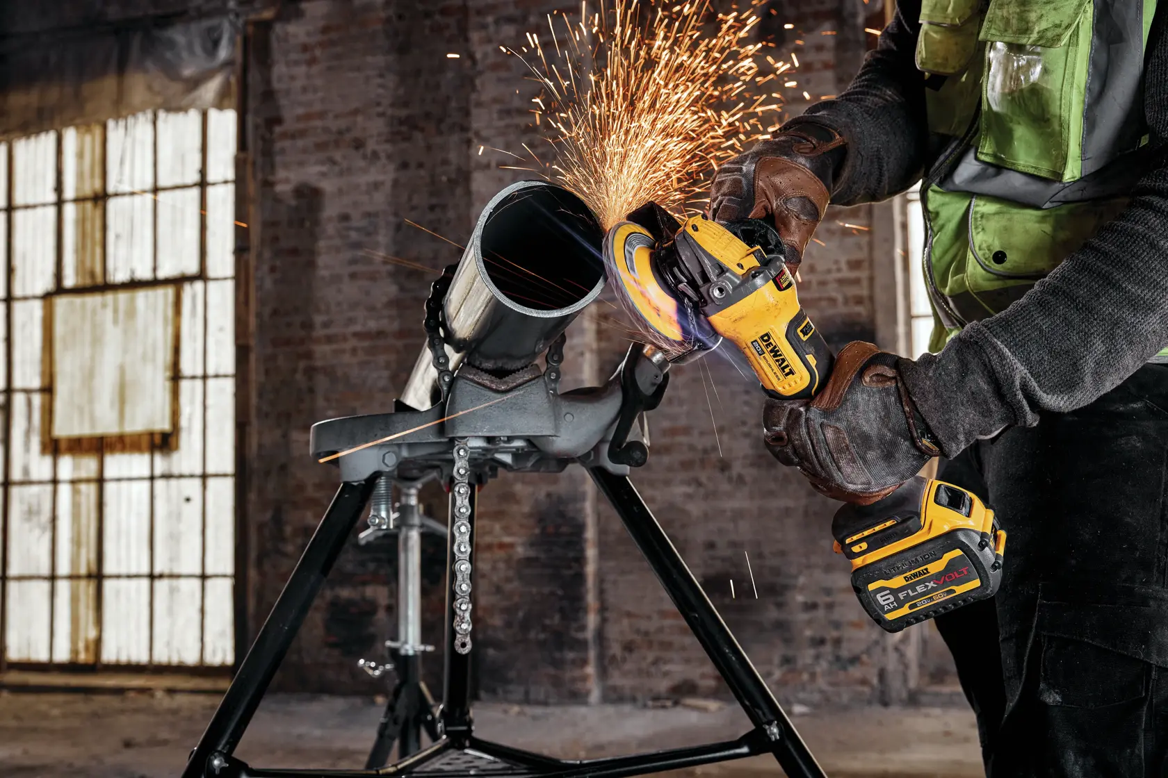 A person wearing gloves and a work vest operates a DEWALT cordless angle grinder, producing sparks while cutting a metal pipe secured on a stand inside an industrial setting.