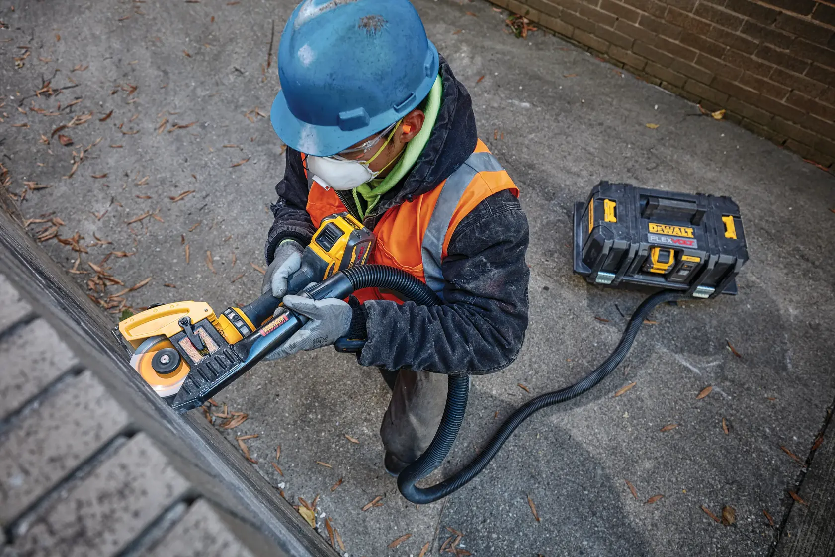 A worker wearing a blue hard hat, orange safety vest, and gloves is using a DEWALT power tool to grind or cut masonry. The tool is connected by a hose to a DEWALT dust extractor placed on the ground nearby. The worker's face is blurred for privacy.