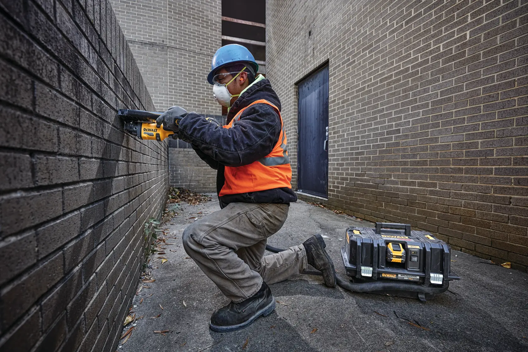 A person wearing a hard hat, safety vest, gloves, and work boots is kneeling while using a DEWALT power tool on a brick wall. A DEWALT tool case with equipment is placed on the ground nearby. The scene takes place outdoors between two brick buildings.