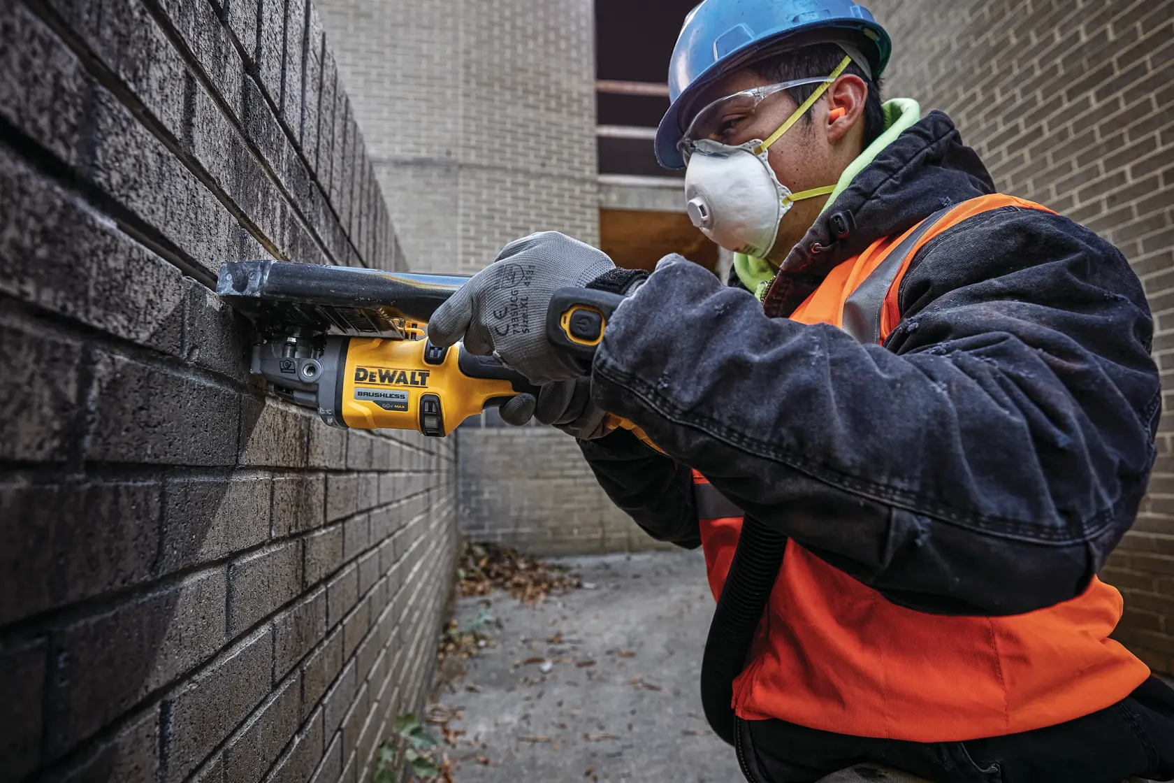 A person wearing a blue hard hat, safety gloves, and an orange safety vest is using a yellow DEWALT cordless power tool to cut through mortar joints on a brick wall. The scene takes place outdoors in a construction setting.