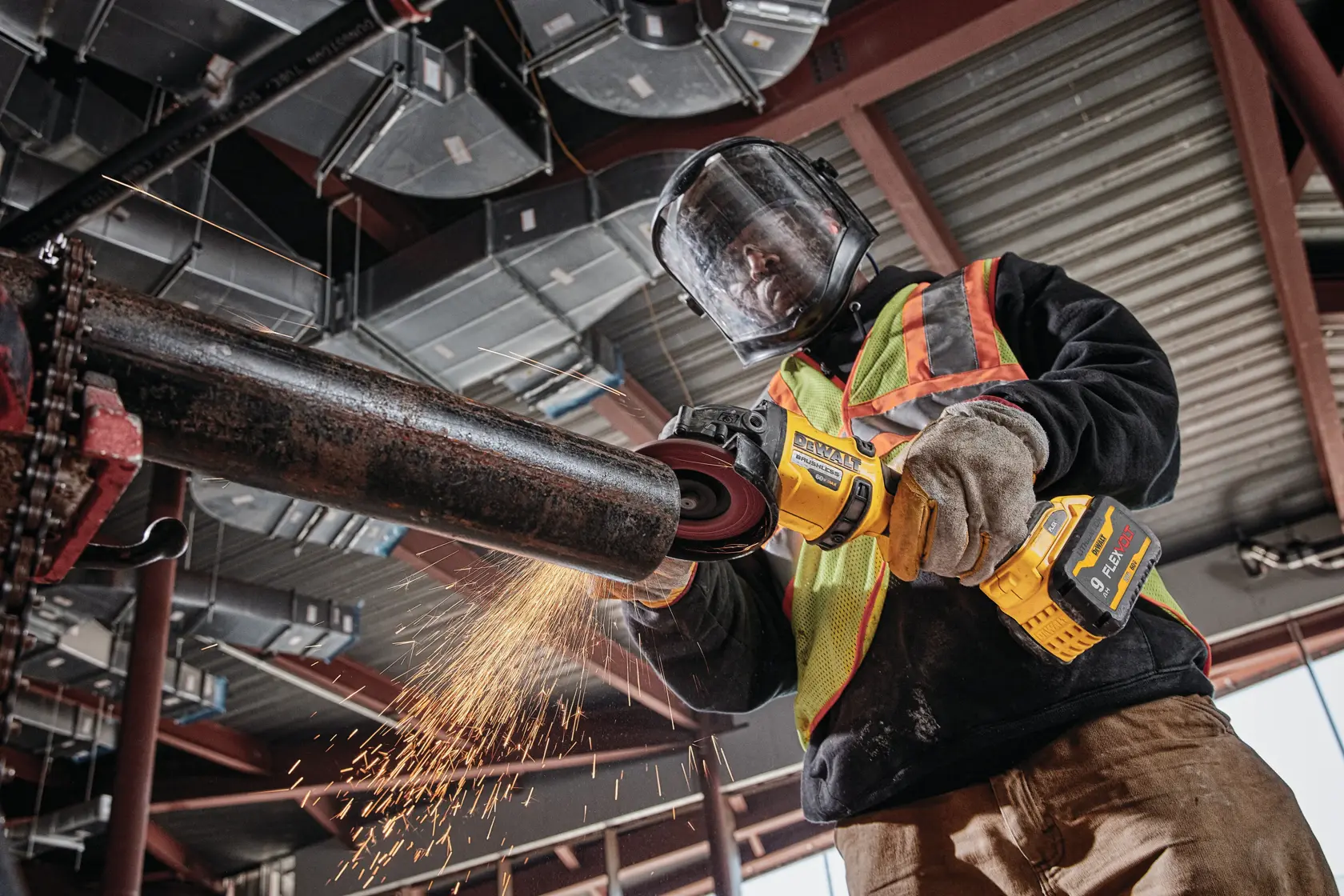 A worker wearing a safety helmet, face shield, gloves, and a hi-vis vest uses a DEWALT cordless angle grinder to cut a metal pipe, producing sparks, inside an industrial building.
