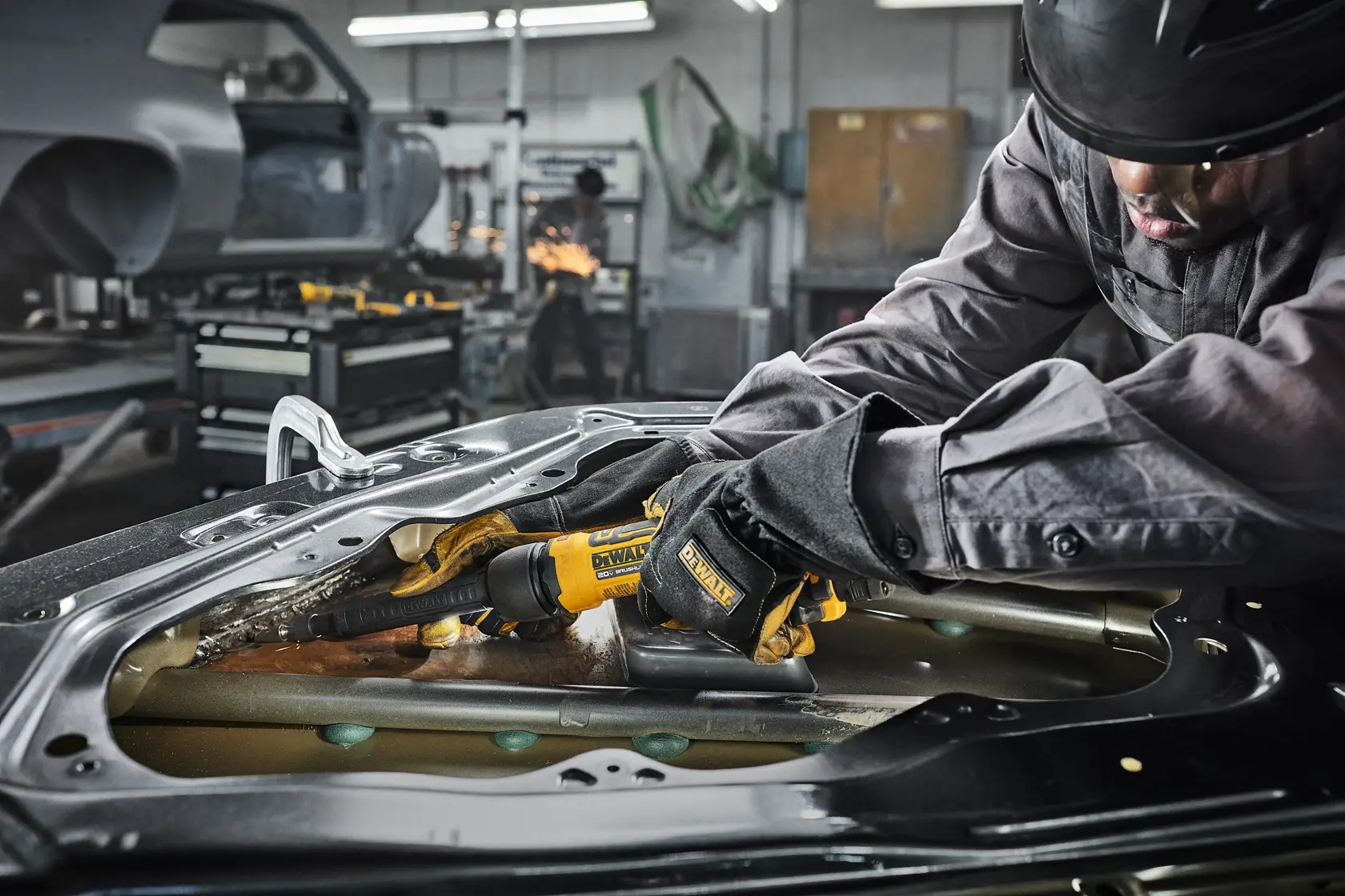 A person wearing protective gloves and a helmet is using a DEWALT power tool to grind or cut metal inside a vehicle body part in an industrial workshop. The environment includes other vehicle parts, equipment, and machinery in the background.