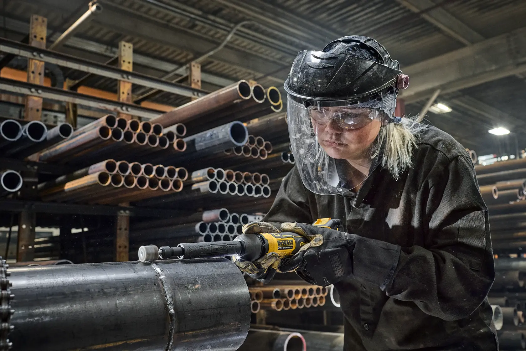 A person wearing protective gloves and a helmet is using a DEWALT power tool to grind or cut metal inside a vehicle body part in an industrial workshop. The environment includes other vehicle parts, equipment, and machinery in the background.