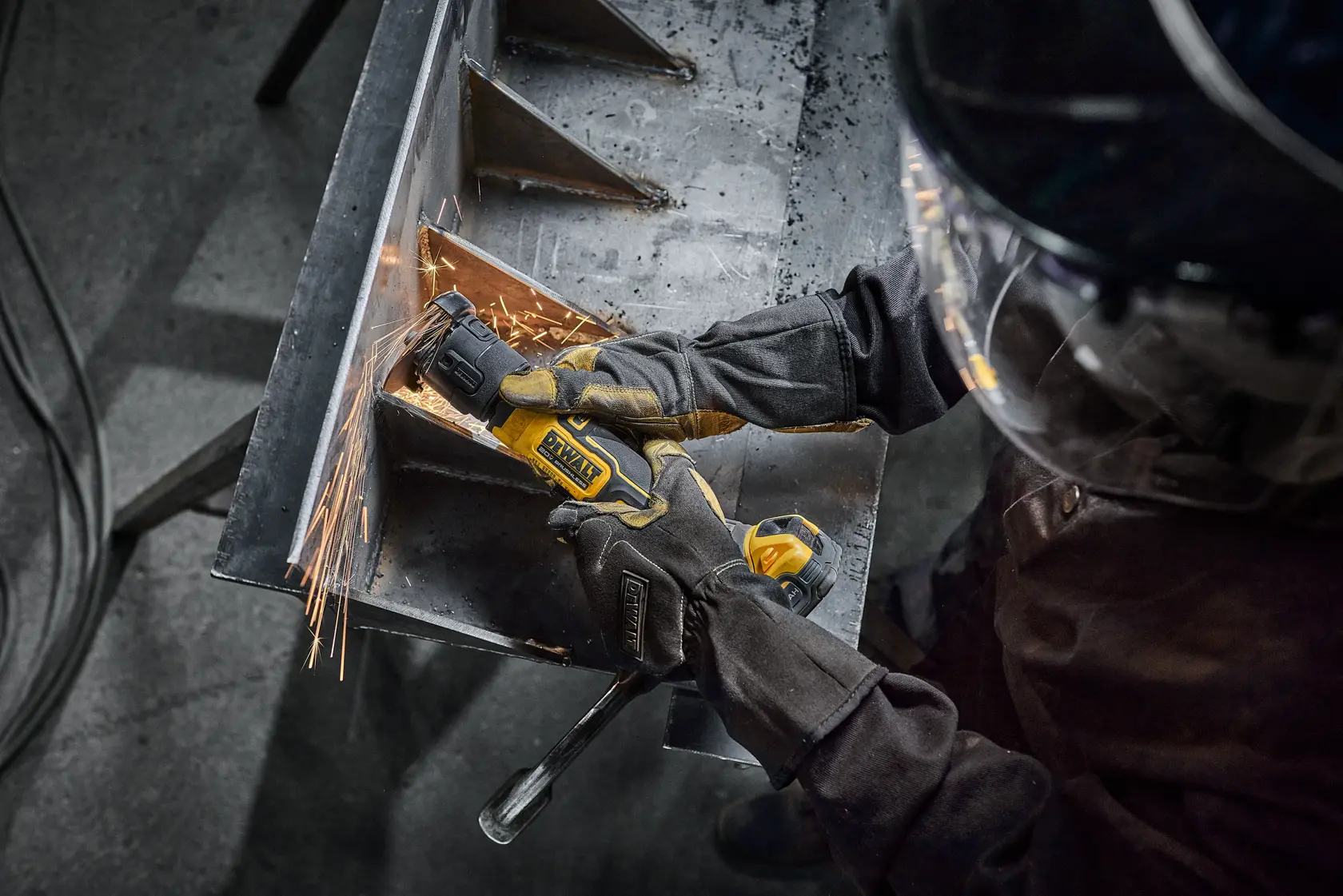 A person wearing protective gloves and a face shield uses a DEWALT cordless angle grinder to grind or cut metal, producing sparks. The tool is being used on a metal workpiece in a workshop setting.