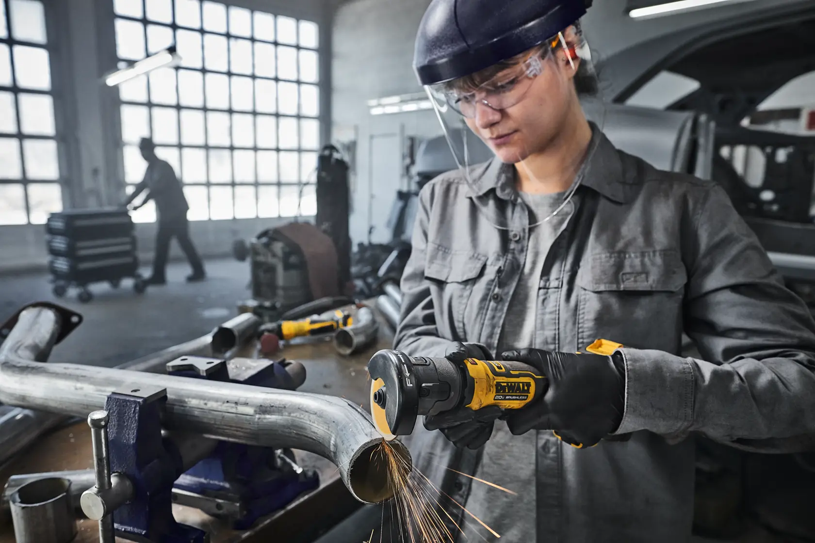 A person using a DEWALT cordless angle grinder to cut a metal pipe in a workshop. Sparks are visible from the cutting process. The individual is wearing protective gloves and a helmet. The background shows workshop equipment and another person working.