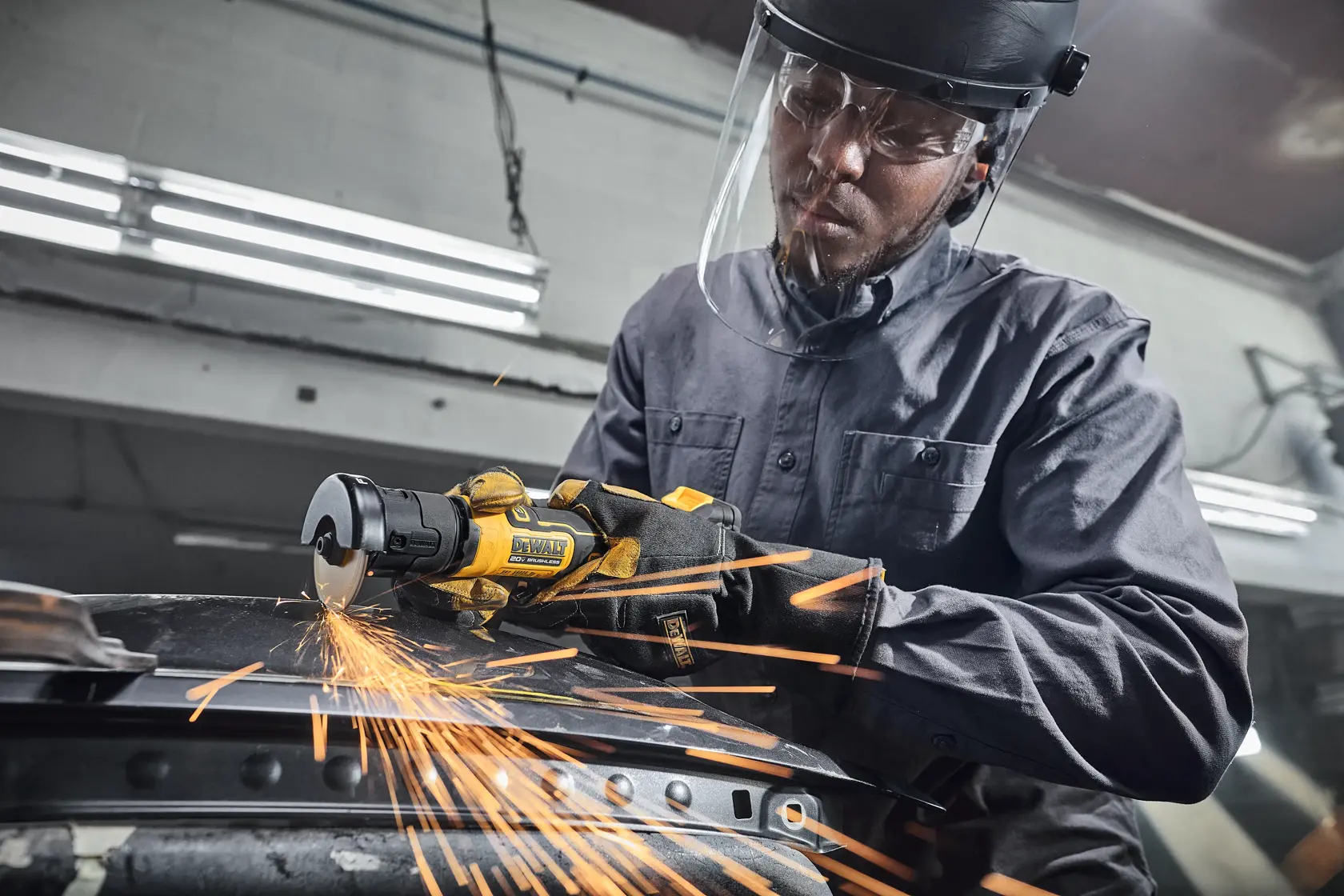 A person using a DEWALT cordless angle grinder to cut a metal pipe in a workshop. Sparks are visible from the cutting process. The individual is wearing protective gloves and a helmet. The background shows workshop equipment and another person working.