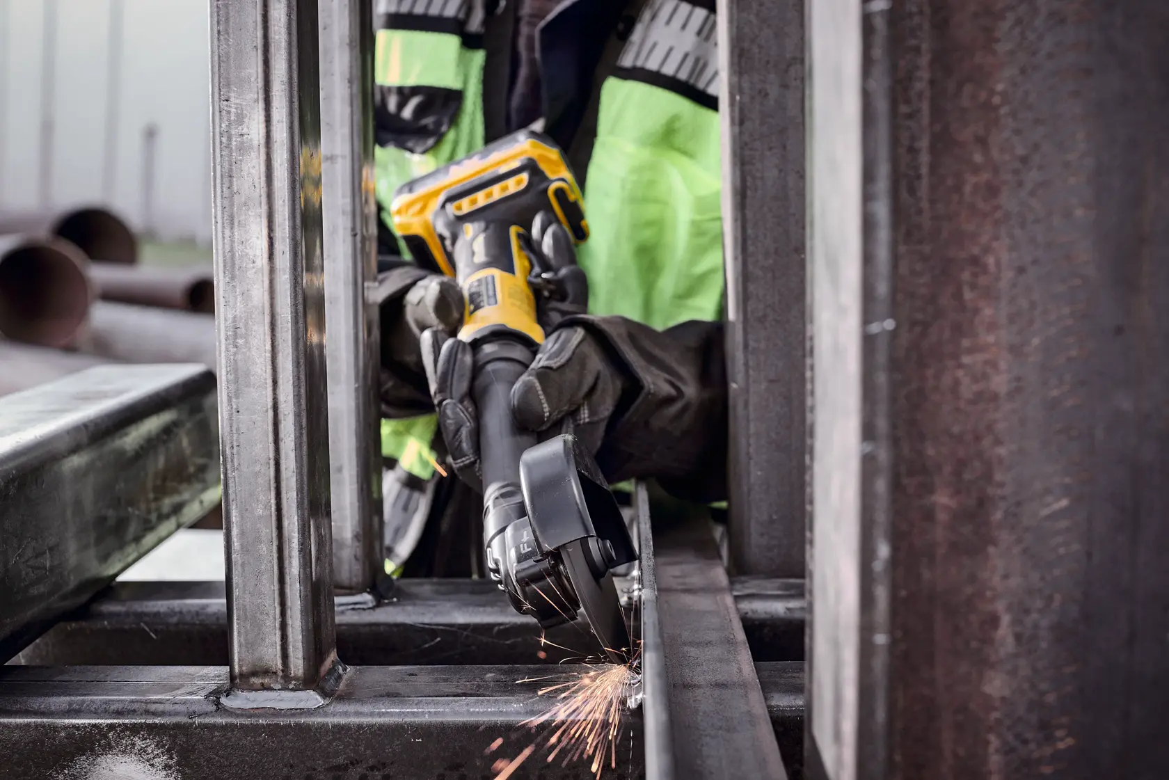 A person wearing protective gloves and a high-visibility jacket uses a DEWALT cordless angle grinder to cut or grind a metal beam, with sparks flying during the process.
