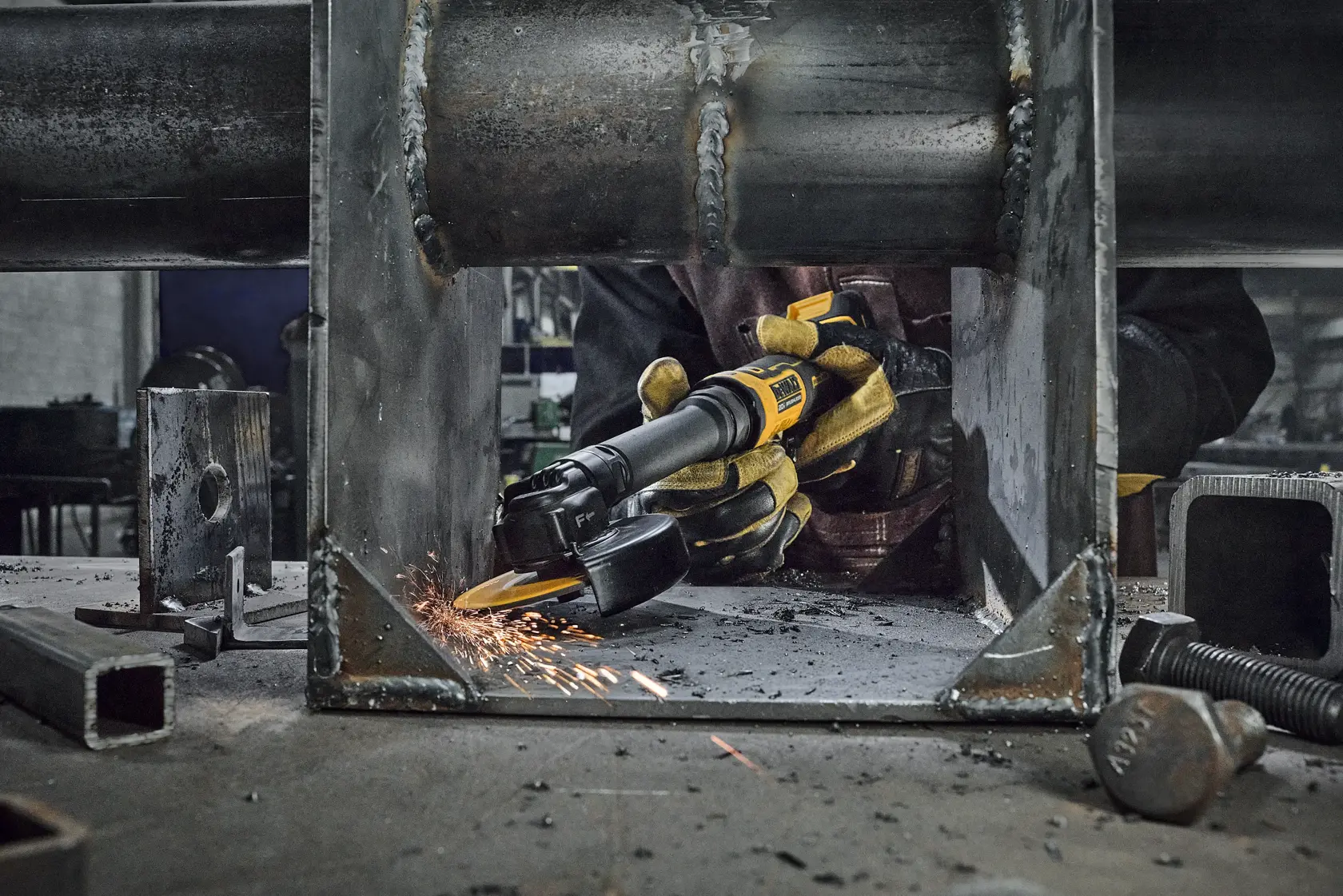 A person wearing protective gloves uses a yellow DEWALT cordless grinder to smooth the inside of a metal structure, producing sparks in an industrial workshop setting.