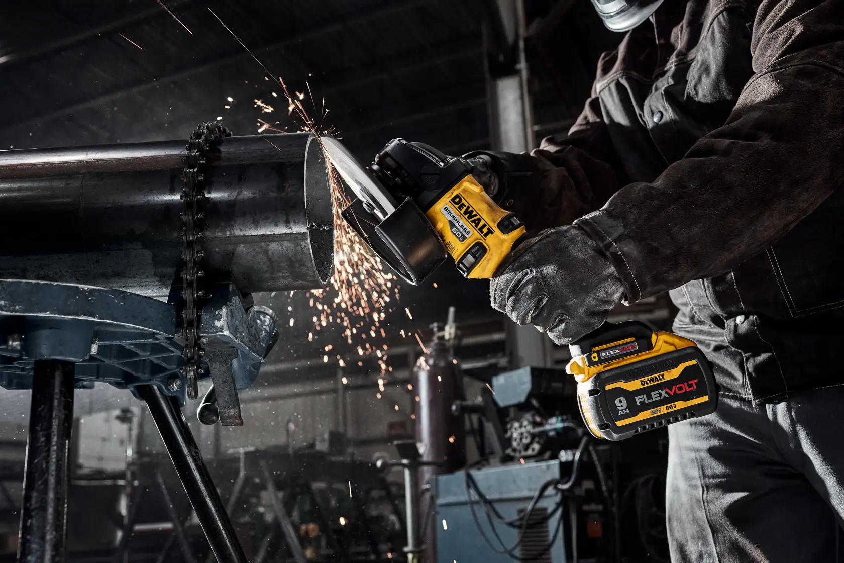 A person wearing protective gear uses a DEWALT cordless angle grinder to cut through a large metal pipe in an industrial workshop, with sparks flying from the cutting process.