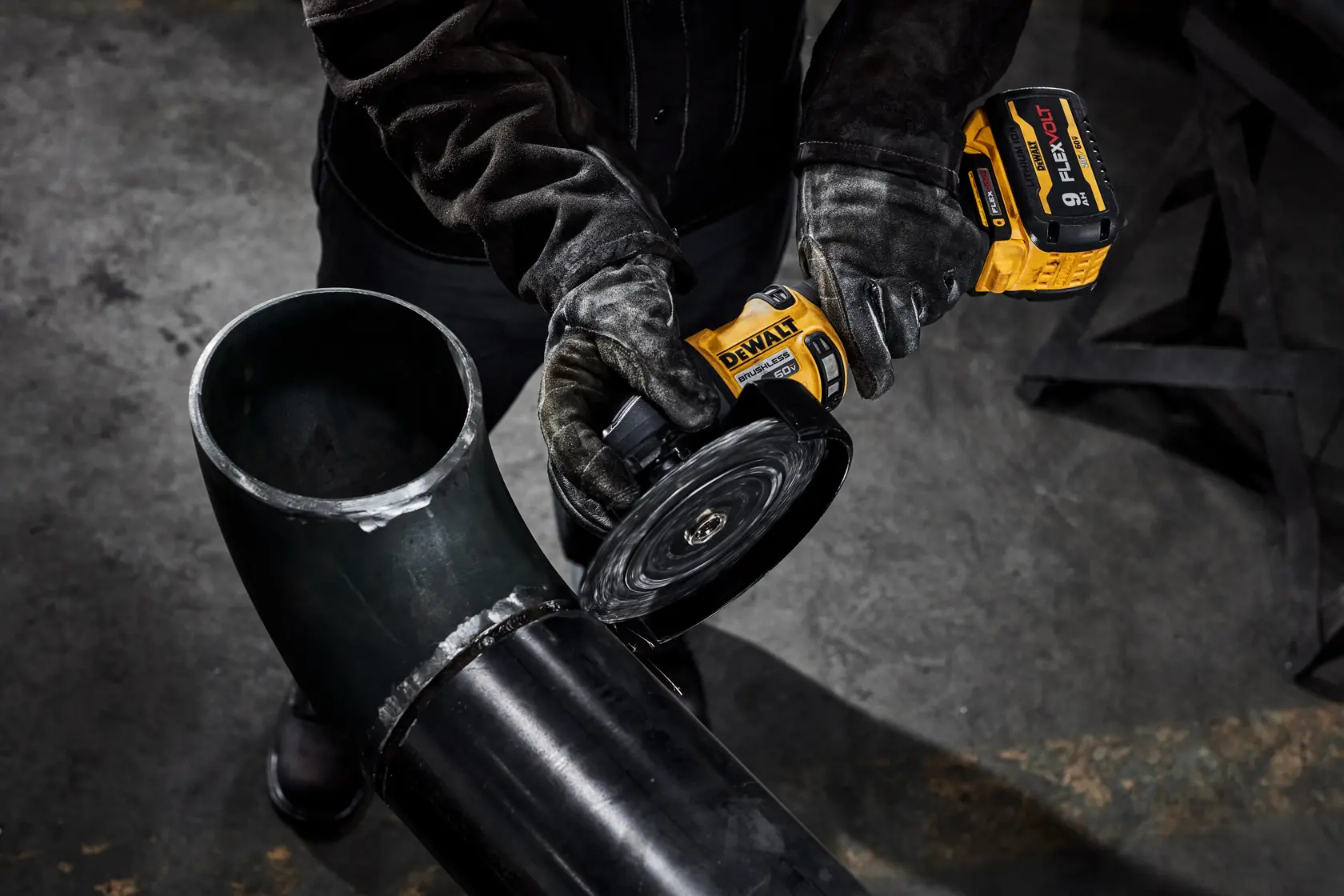 A person wearing protective gloves is using a DEWALT cordless grinder to cut or polish a large black metal pipe in an industrial setting.