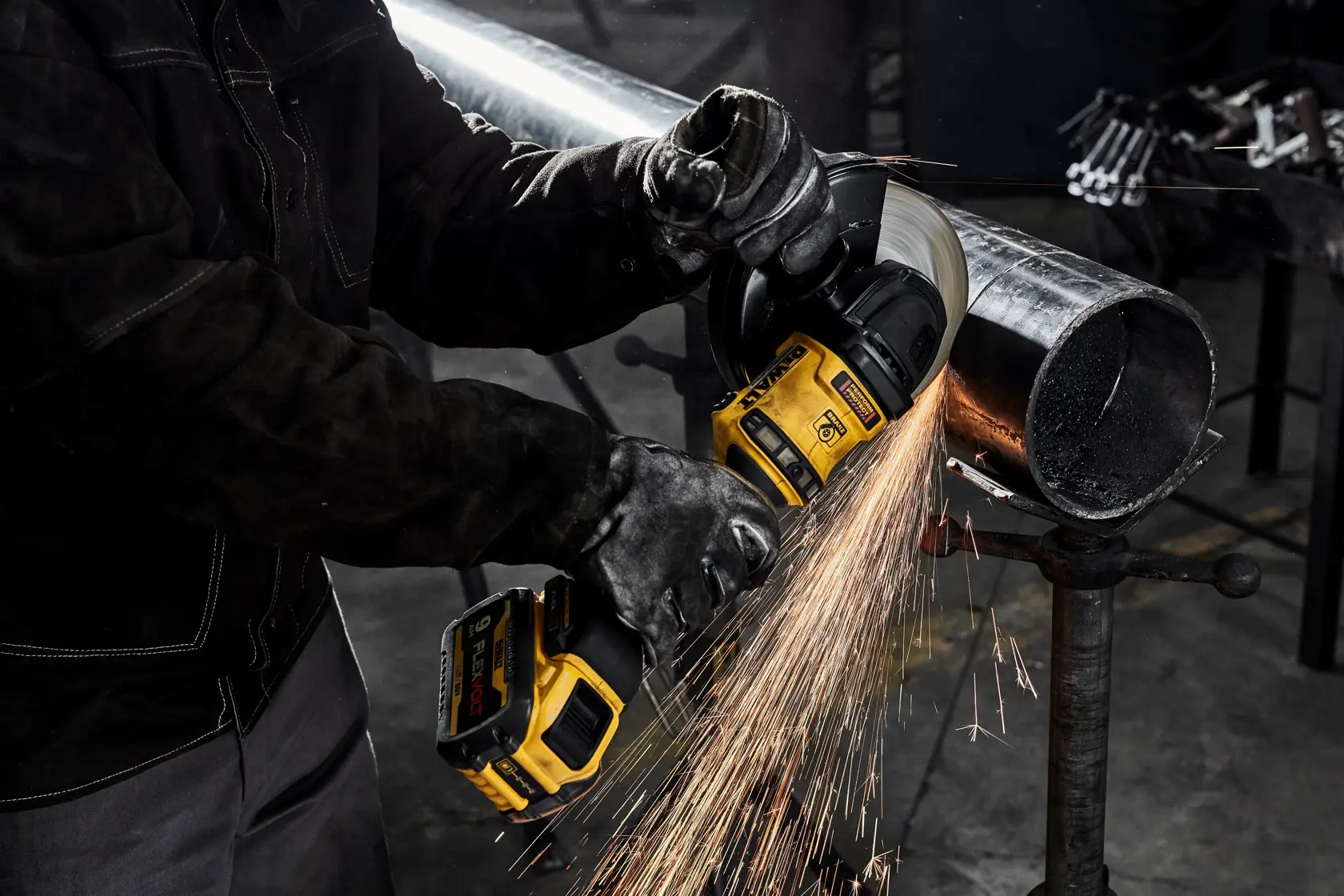 A worker wearing protective gloves uses a DEWALT cordless angle grinder to cut through a metal pipe, producing sparks.