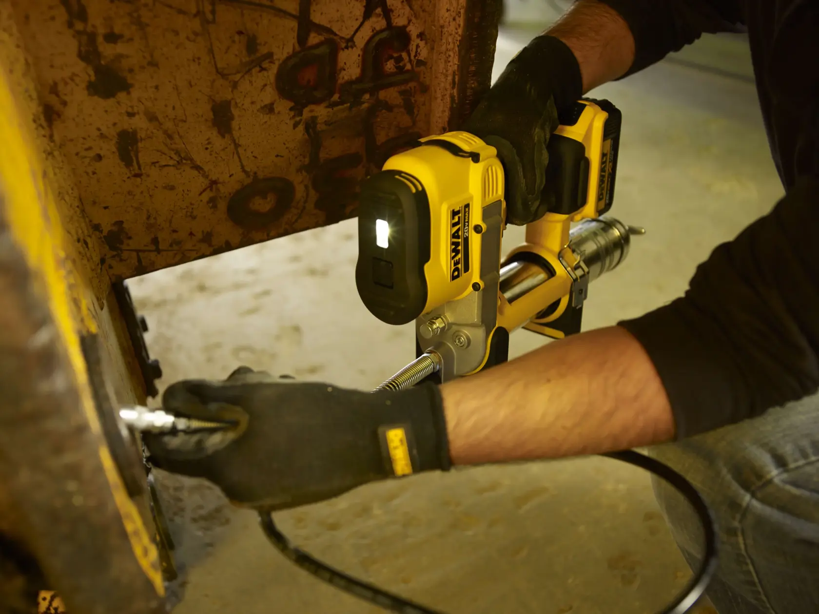 A person wearing gloves uses a DEWALT DCGG571M1 cordless grease gun to lubricate a metal machinery part. The tool is yellow and black, with a built-in LED light illuminating the work area.