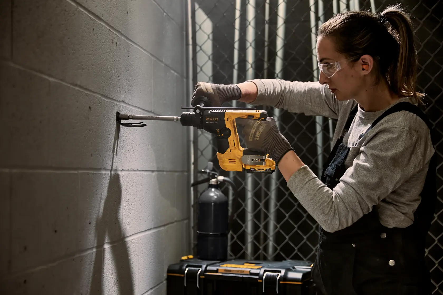 A person using a DEWALT rotary hammer drill to drill into a concrete wall. The individual is wearing gloves and work overalls, and their face is blurred. There is a tool case and other equipment in the background.