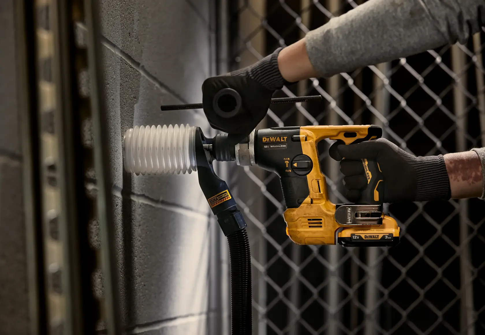 A person wearing black gloves is using a DEWALT cordless rotary hammer drill to drill into a concrete wall. The tool is yellow and black, and is attached to a dust extraction hose with a flexible white dust collection attachment.