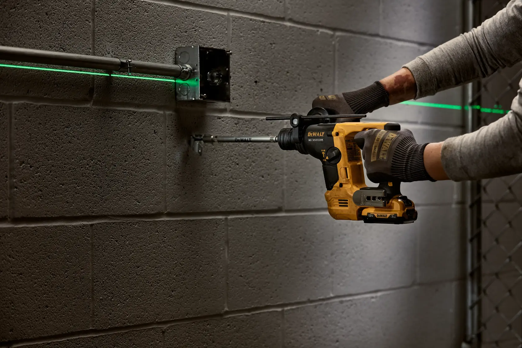 A person wearing gloves uses a DEWALT cordless rotary hammer drill to drill into a concrete wall. Green laser lines are visible, marking the placement of metal conduit pipes and an electrical box.