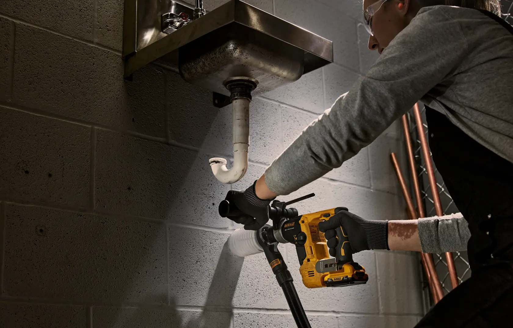 A person using a DEWALT cordless rotary hammer drill to make a hole in a cinder block wall underneath a metal sink, with a white pipe attached. The tool is connected to a dust extraction hose. The person's face is blurred.