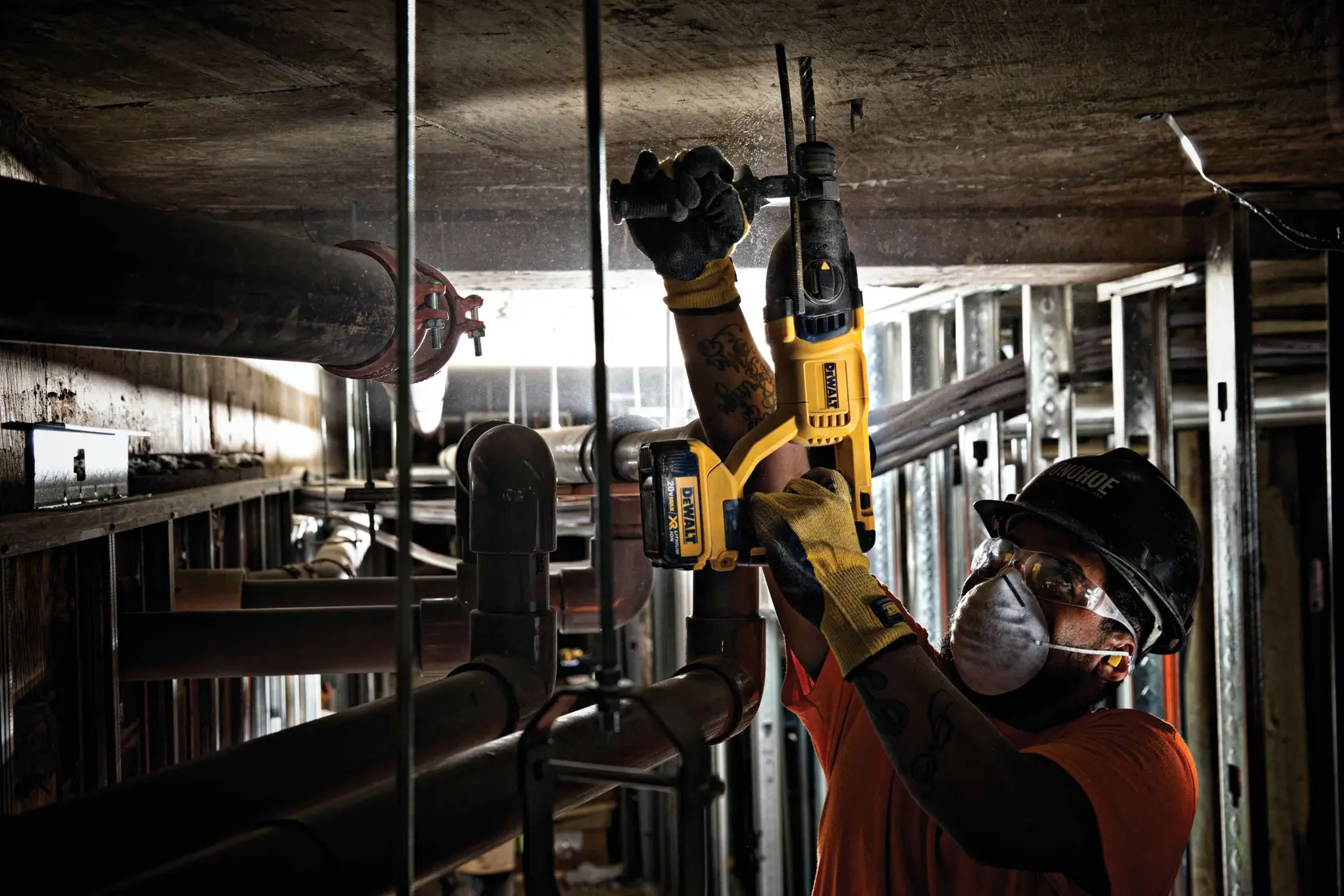 A construction worker wearing gloves and a hard hat is using a yellow DEWALT cordless rotary hammer drill to work on a concrete ceiling in an industrial setting with exposed pipes and metal framing.
