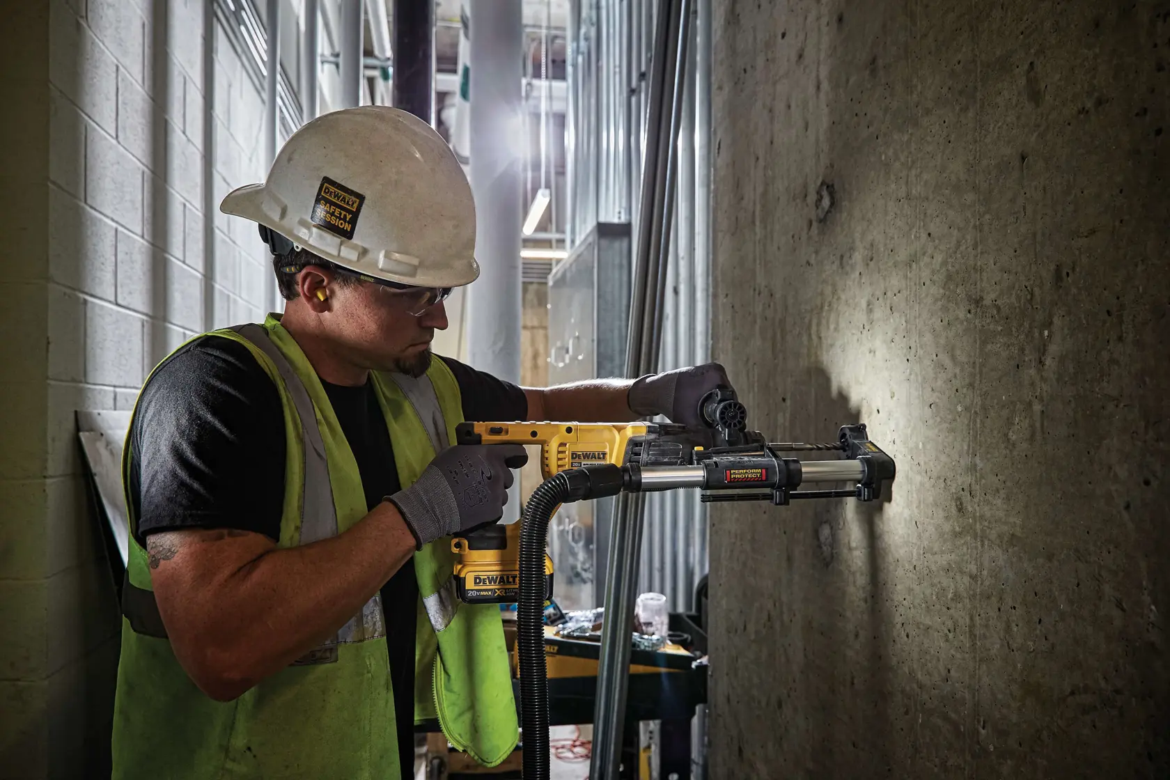 A person wearing a safety helmet, gloves, and a reflective vest is using a yellow DEWALT rotary hammer drill (SKU: DCH133B) to drill into a concrete wall inside a construction site. The drill is attached to a dust extraction system for cleaner operation. The person's face is blurred.