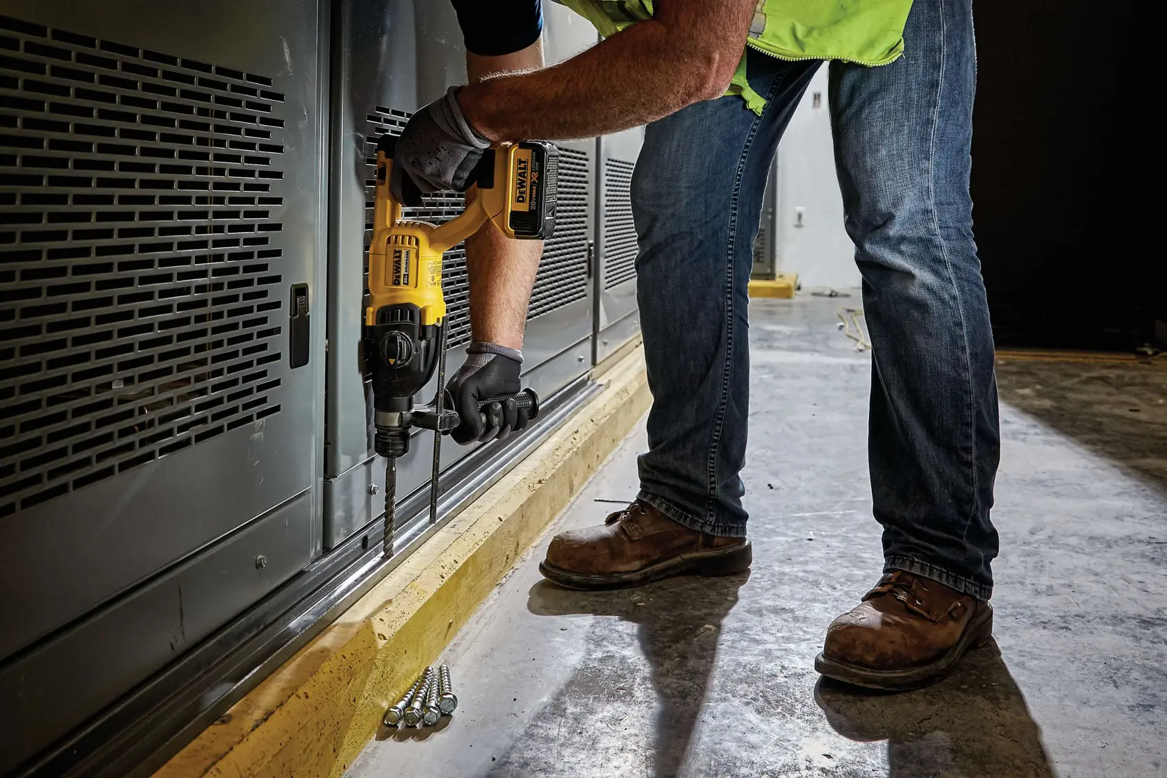 A person wearing jeans, work boots, and gloves uses a DEWALT cordless rotary hammer drill to drill into a metal surface near the floor in an industrial or construction setting.