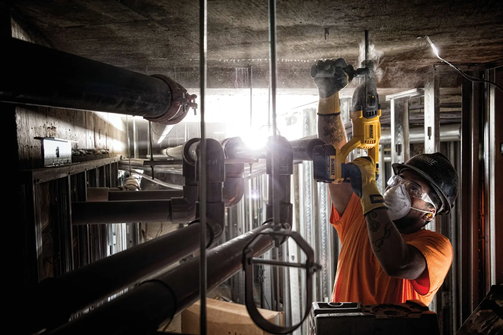 A worker wearing protective gear uses a yellow DEWALT cordless rotary hammer drill (model DCH133B) to drill into a concrete ceiling in an industrial setting with metal pipes and construction materials visible.