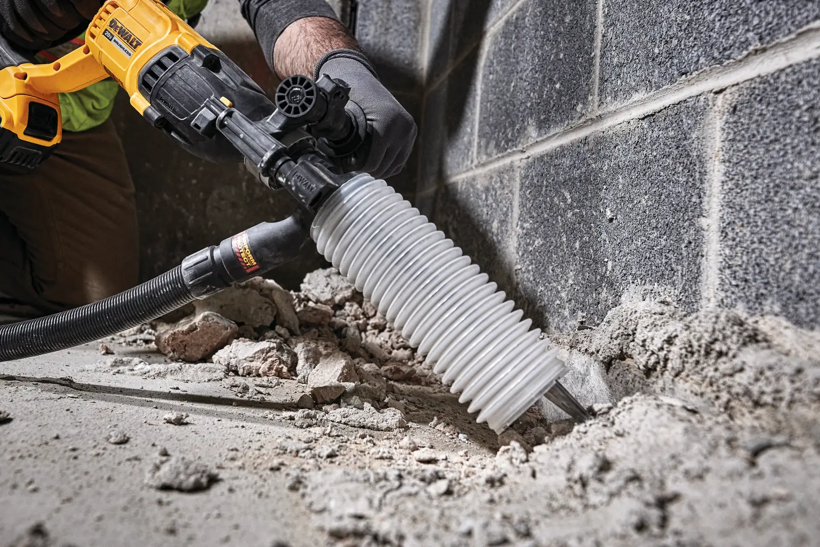 A person using a DEWALT rotary hammer drill with a dust collection attachment to break concrete near a wall. The tool is yellow and black, and debris is visible on the ground.