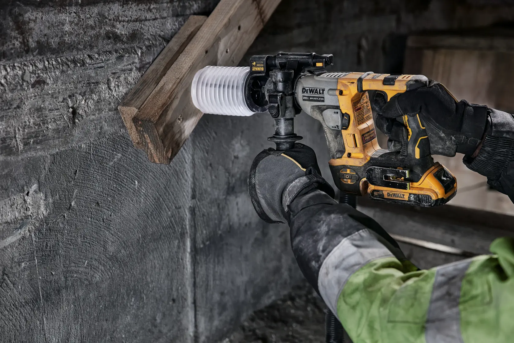 A person using a DEWALT cordless rotary hammer drill to bore a hole through a wooden beam and concrete wall. The user is wearing gloves and a work jacket.