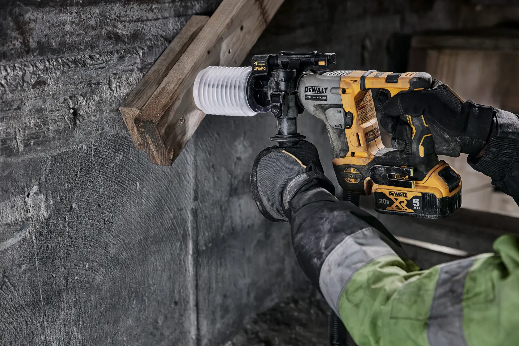 A person using a DEWALT cordless rotary hammer drill (model DCH172B) to drill into a piece of wood attached to a concrete wall. The drill features a yellow and black design and a 20V lithium-ion battery. The individual is wearing gloves and a green work jacket.