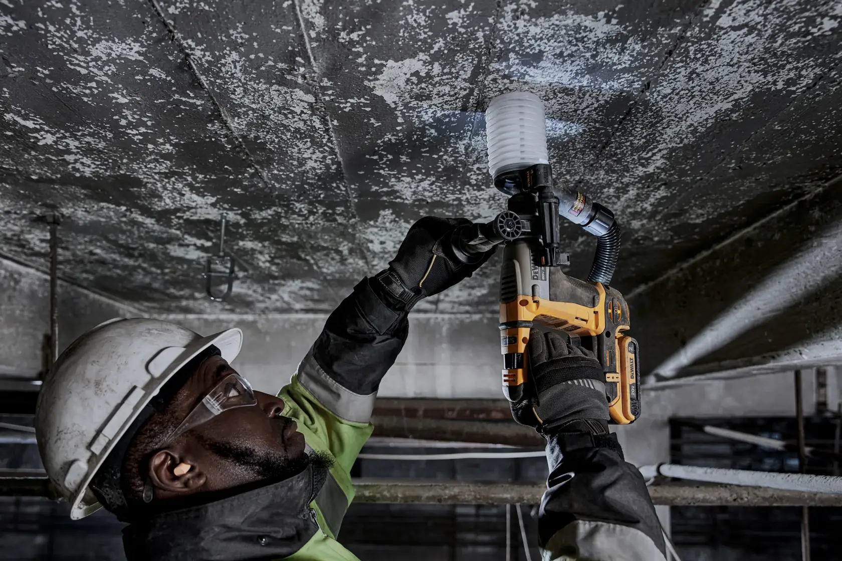 A construction worker wearing safety gear uses a DEWALT cordless rotary hammer drill (model DCH172D2) to drill into a concrete ceiling. The worker's face is obscured for privacy.