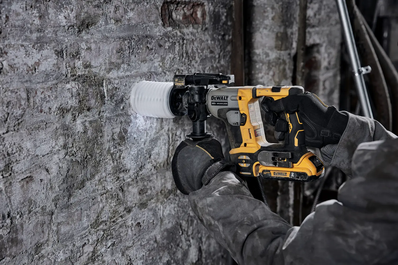 A person wearing work gloves uses a DEWALT cordless rotary hammer drill with a dust extraction attachment to drill into a concrete wall.
