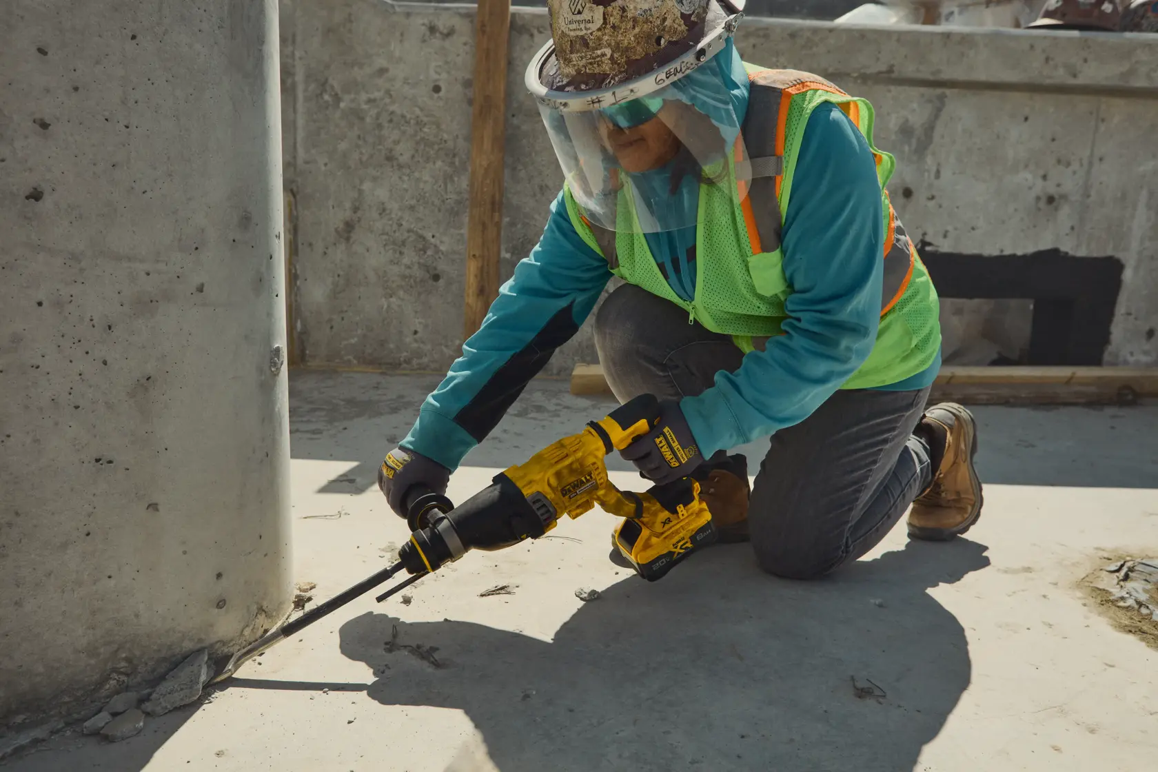 A construction worker wearing safety gear uses a yellow DEWALT rotary hammer drill (model DCH263B) to chip concrete at the base of a pillar on a construction site.