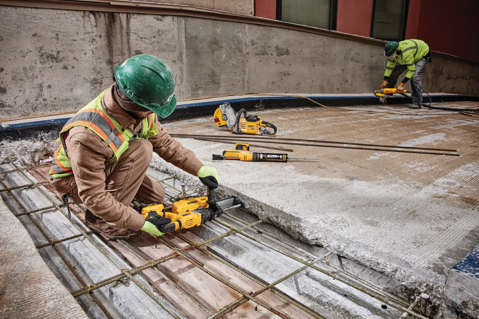 Two construction workers wearing safety gear are using DEWALT power tools to work on a concrete structure with exposed rebar at a construction site.