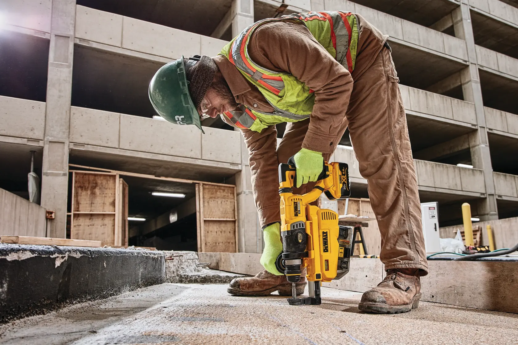 A construction worker wearing safety gear uses a DEWALT cordless rotary hammer drill on a concrete surface at a building site.