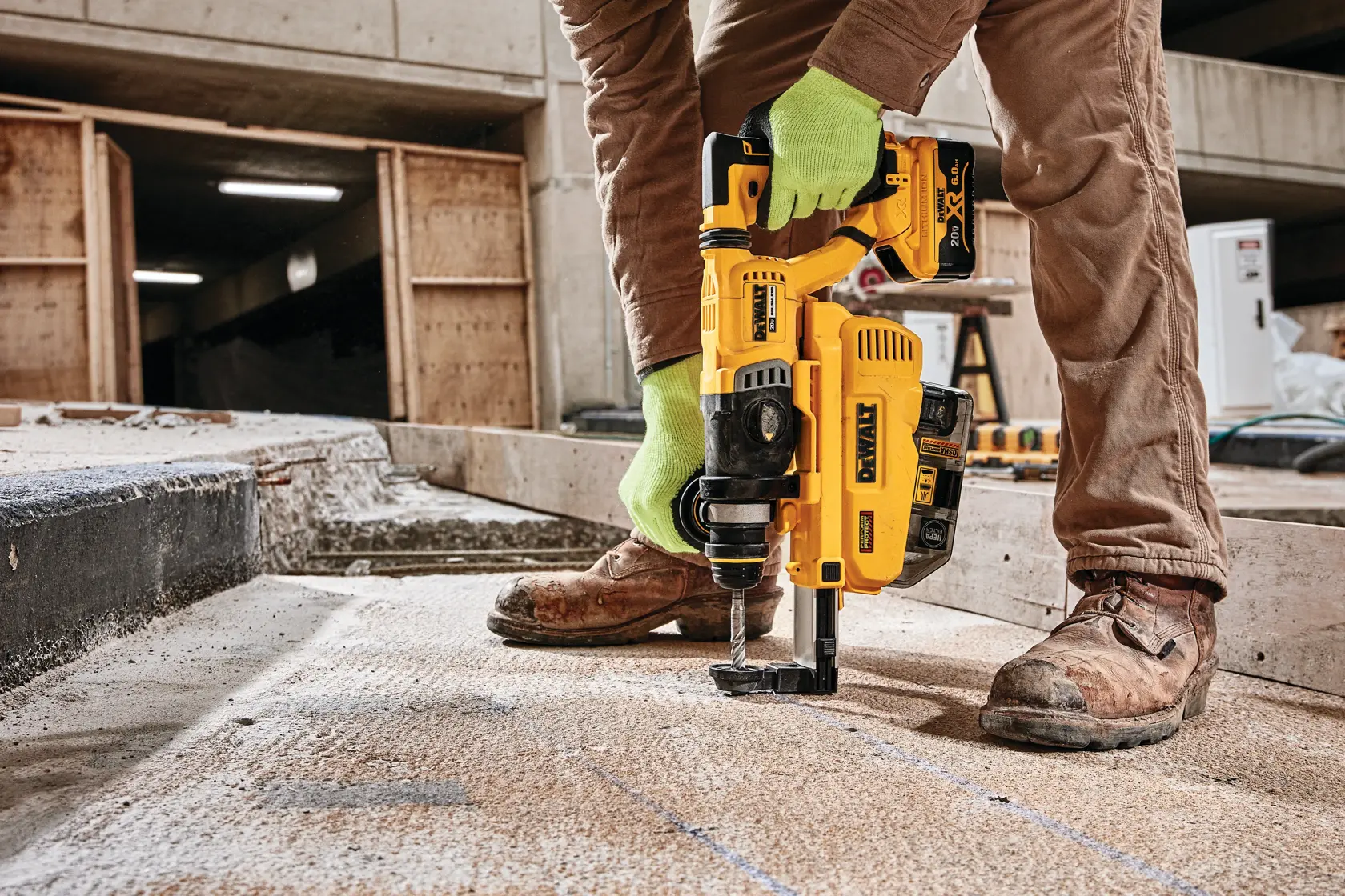A person wearing work gloves and brown pants is using a DEWALT DCH263R2DH rotary hammer drill on a concrete surface at a construction site.