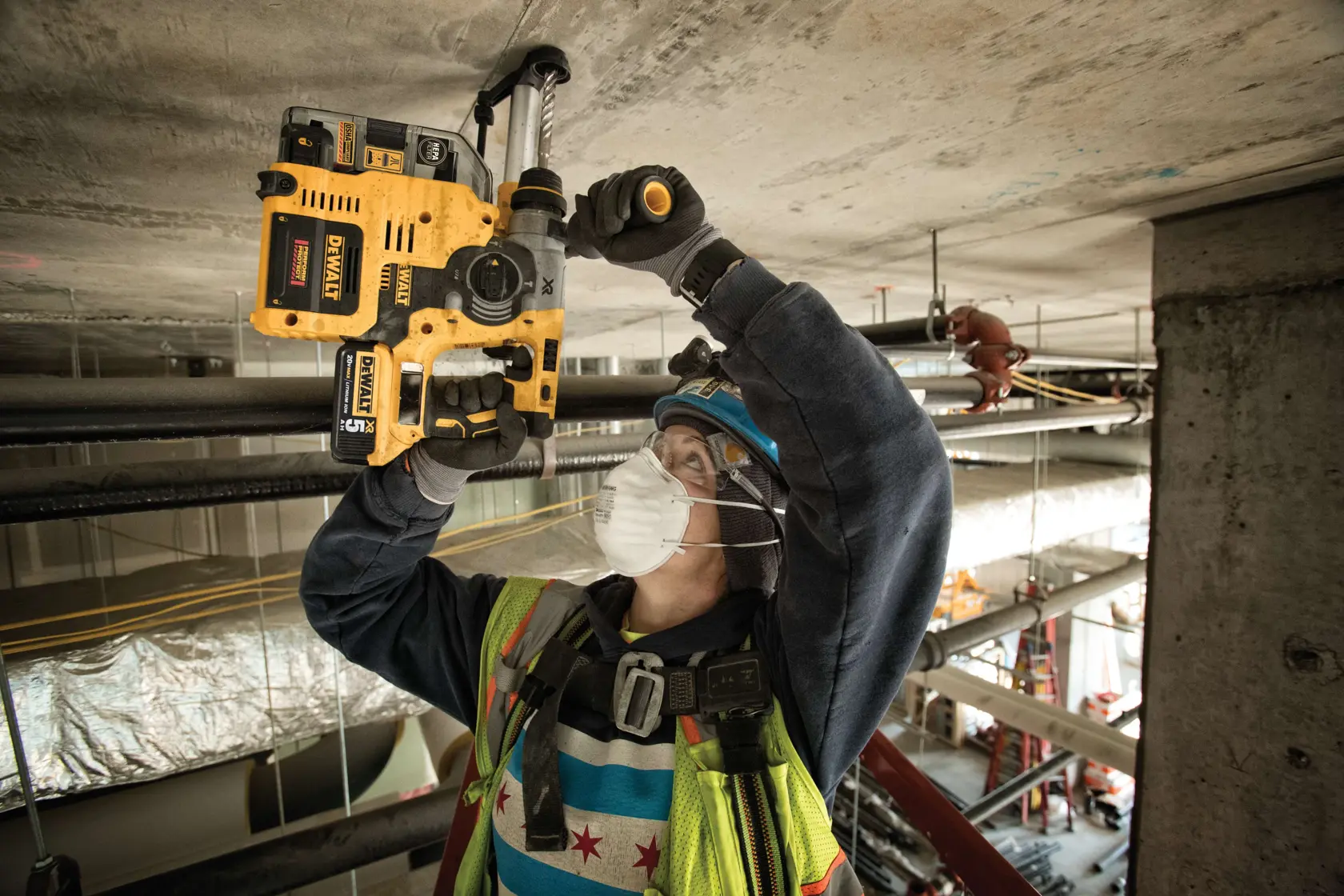 A construction worker wearing a safety vest, hard hat, gloves, and a face mask is using a DEWALT cordless rotary hammer drill to drill into a concrete ceiling at an industrial worksite.