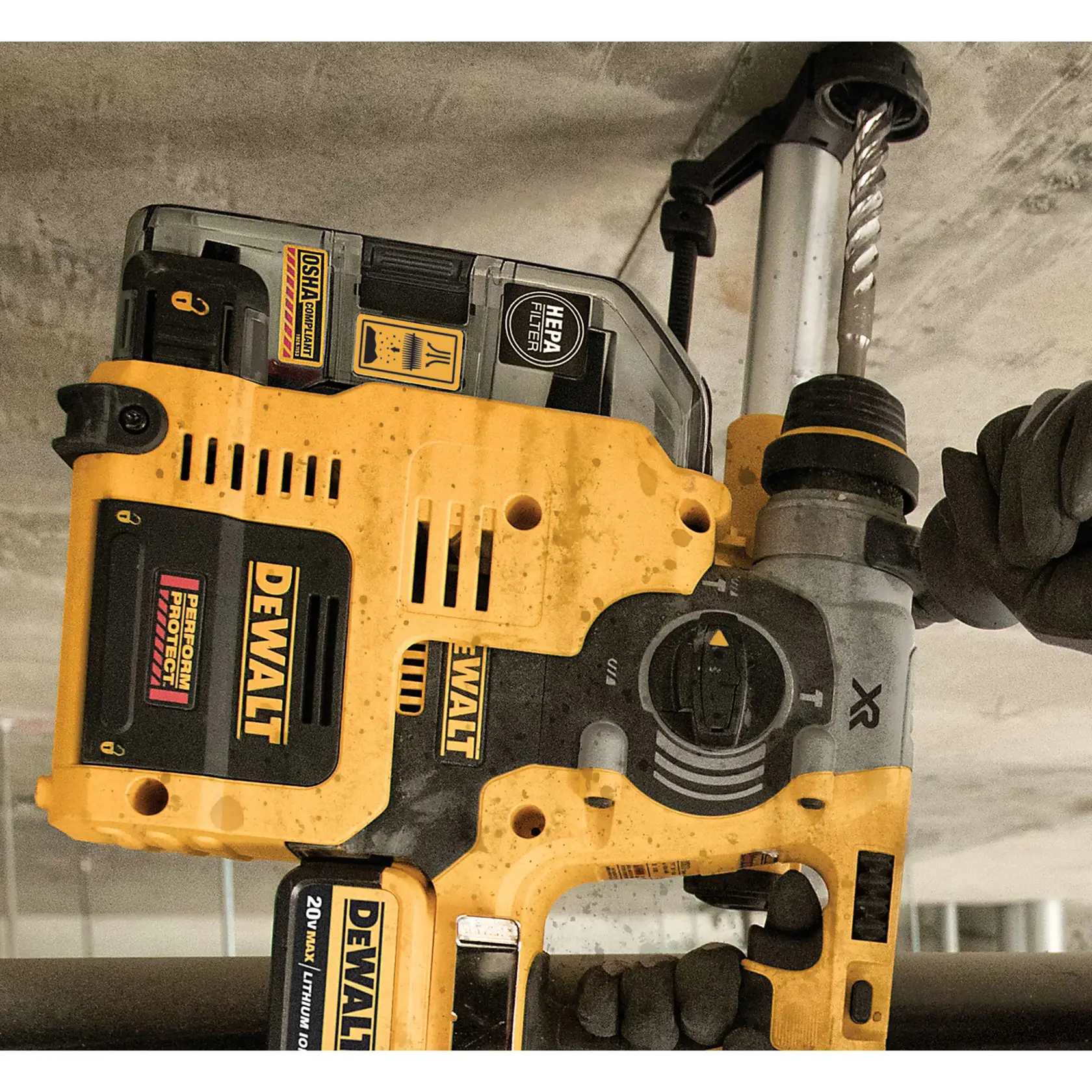 A close-up of a DEWALT cordless rotary hammer drill with dust extraction attachment being used to drill into a concrete ceiling. The HEPA filter and OSHA compliance labels are visible, and a gloved hand is holding the tool.
