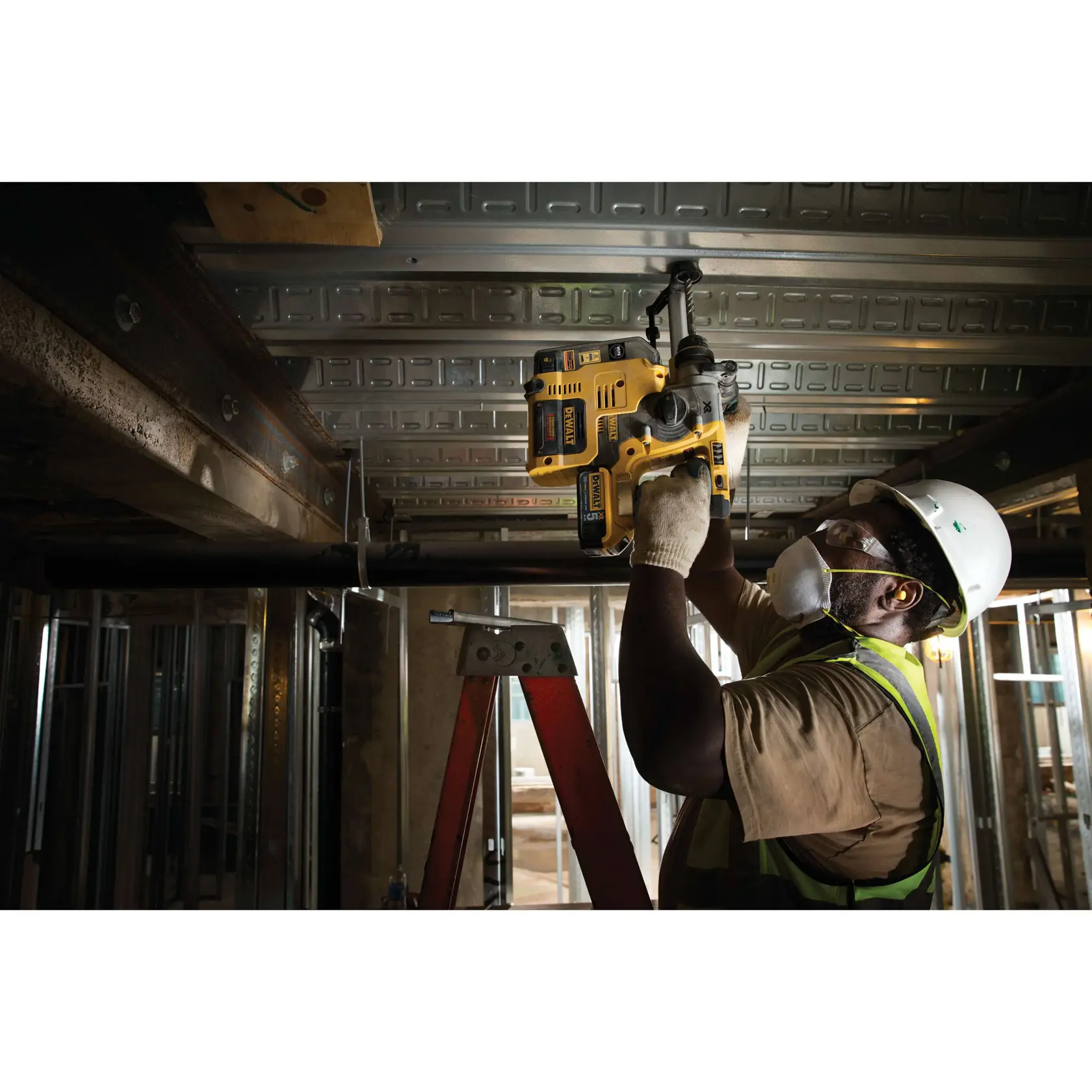 A construction worker wearing a safety helmet, vest, gloves, and face mask is using a DEWALT cordless rotary hammer drill to work on a metal ceiling. The worker is standing on a red ladder inside an industrial building under construction.
