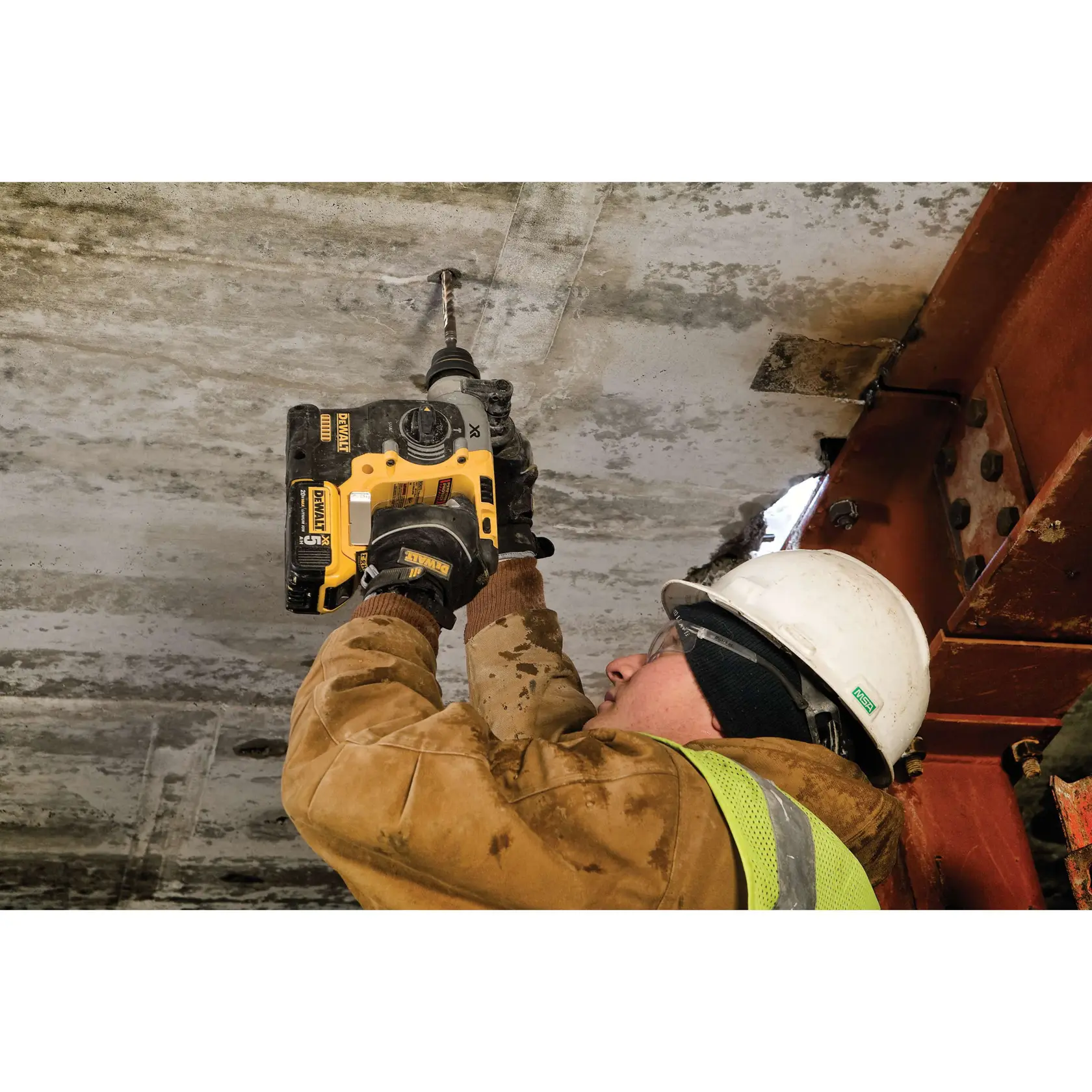 A construction worker wearing a safety helmet and vest uses a DEWALT DCH273P2 rotary hammer drill to drill into a concrete ceiling. The worker is dressed in a brown protective jacket and gloves, and is working on a building structure.