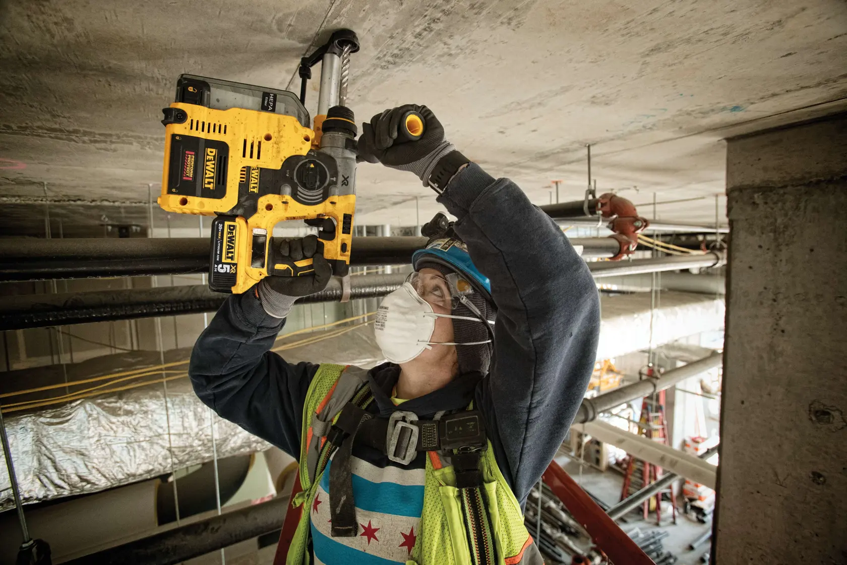 A construction worker wearing a safety vest, hard hat, gloves, and a face mask is using a yellow DEWALT rotary hammer drill to drill into a concrete ceiling at a construction site.