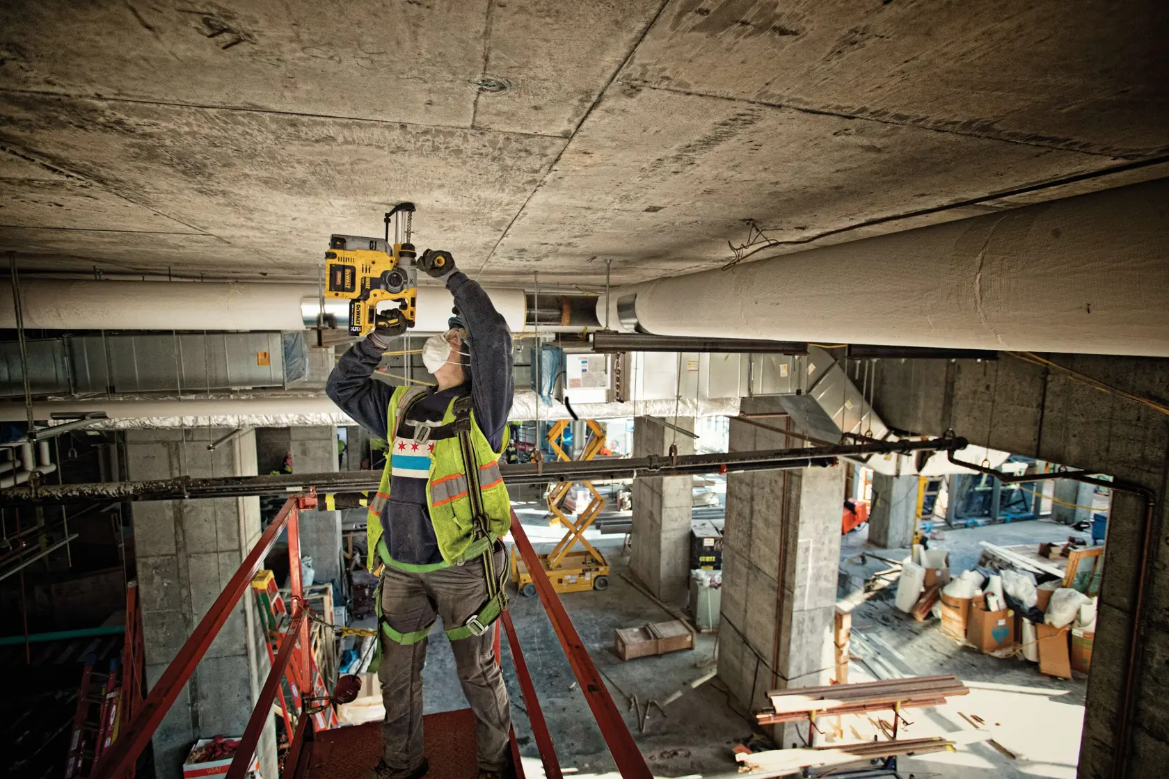 A construction worker wearing a safety vest and face mask is using a DEWALT power tool to drill into a concrete ceiling at an indoor construction site. The worker is standing on a raised platform surrounded by industrial equipment and building materials.